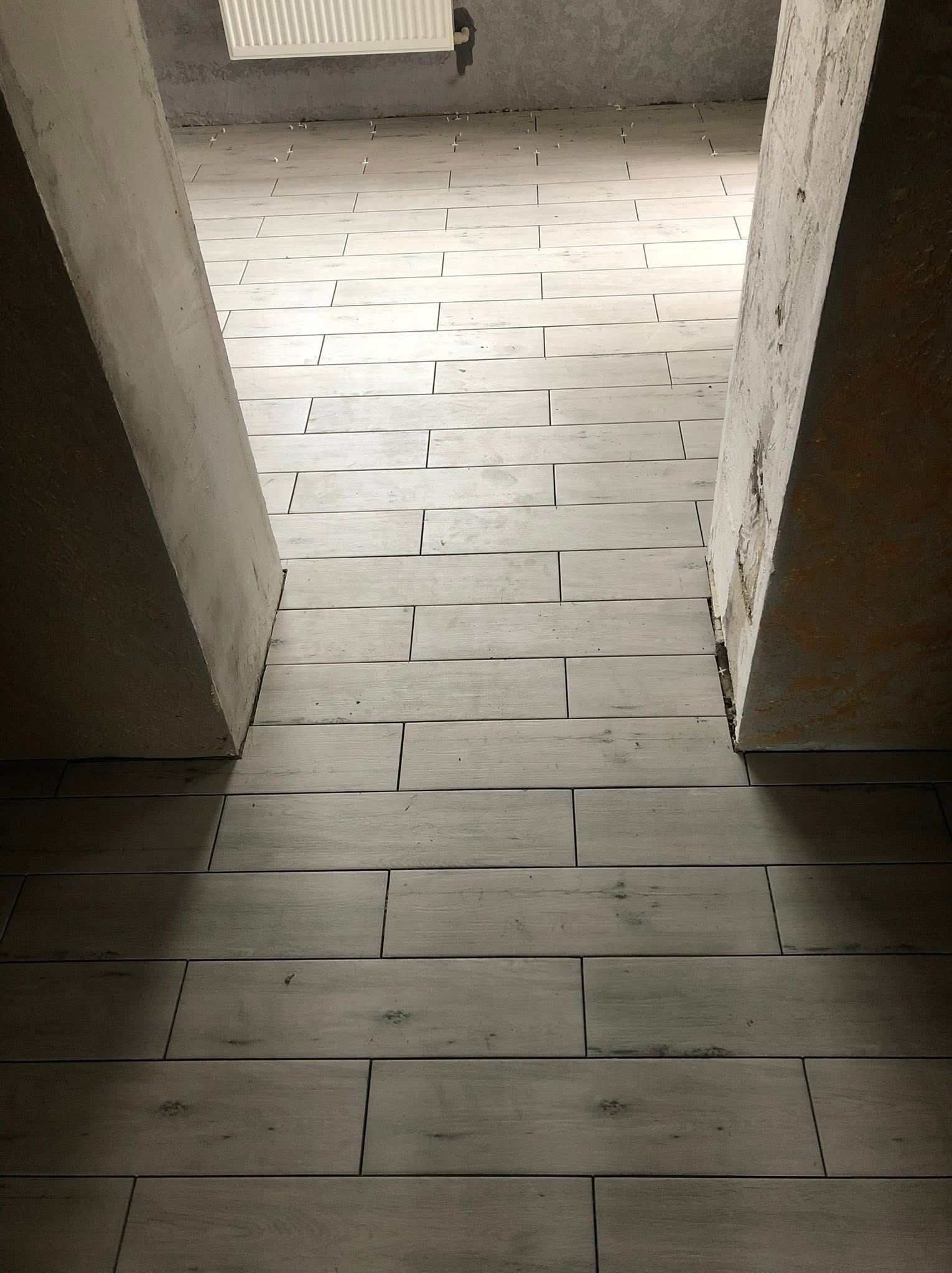 Ułożona podłoga z jasnych płytek imitujących drewno, widoczne krzyżyki dystansowe, surowy beton w przejściu, centralna perspektywa.