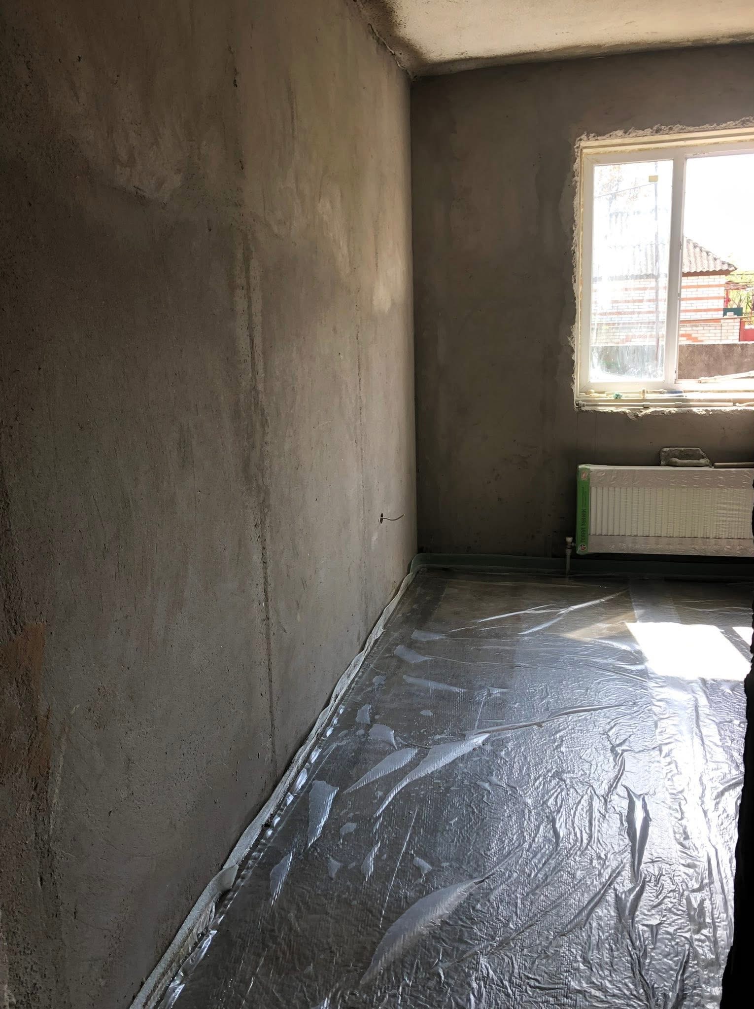 Pokój w surowym stanie: szare ściany po tynkowaniu, okno z grzejnikiem, podłoga zabezpieczona folią. Przygotowanie do dalszych prac wykończeniowych.