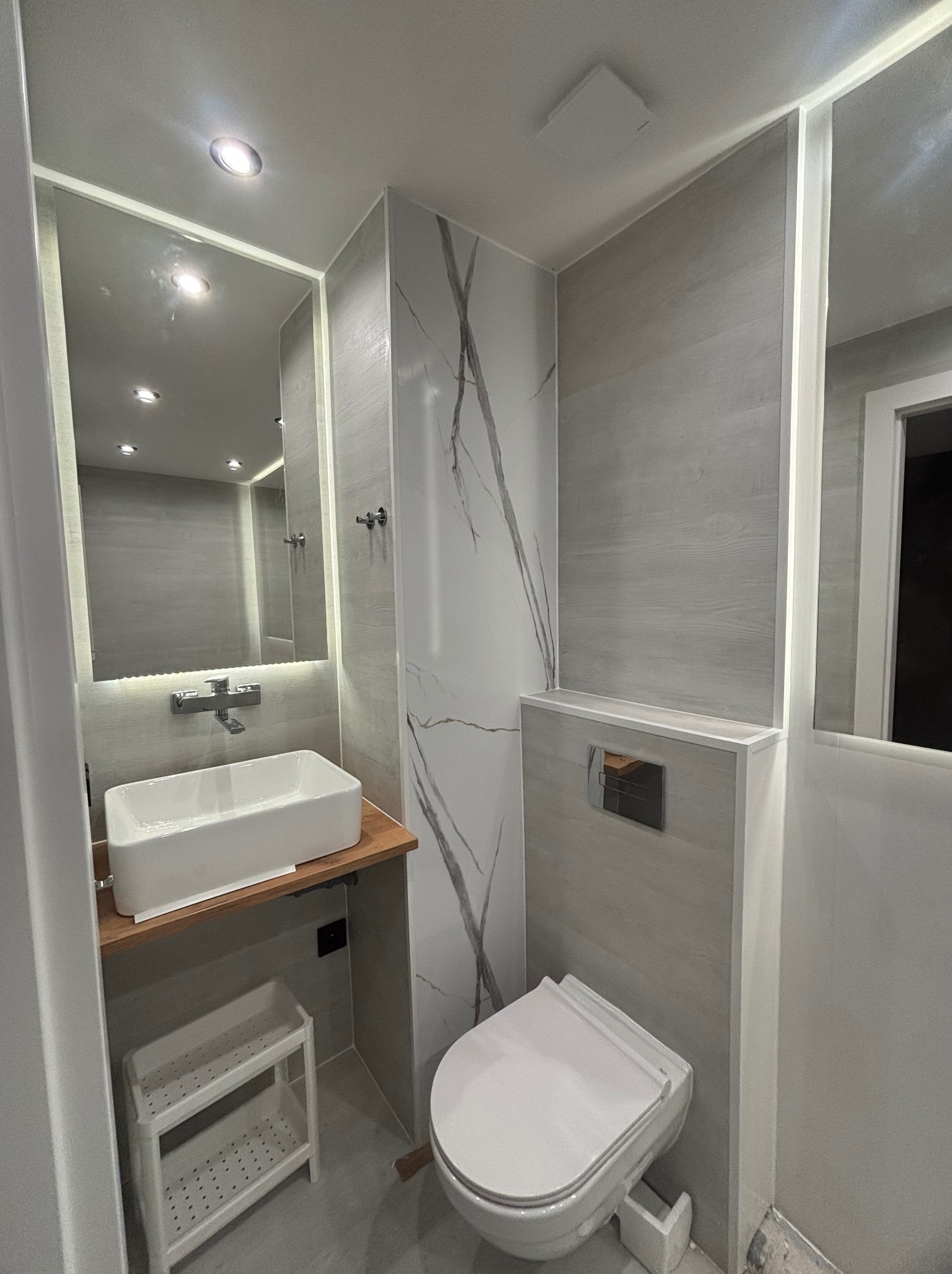 Nowoczesna łazienka po remoncie: biała umywalka na drewnianym blacie, toaleta, szare płytki imitujące drewno, lustro z podświetleniem LED, marmurowe akcenty.