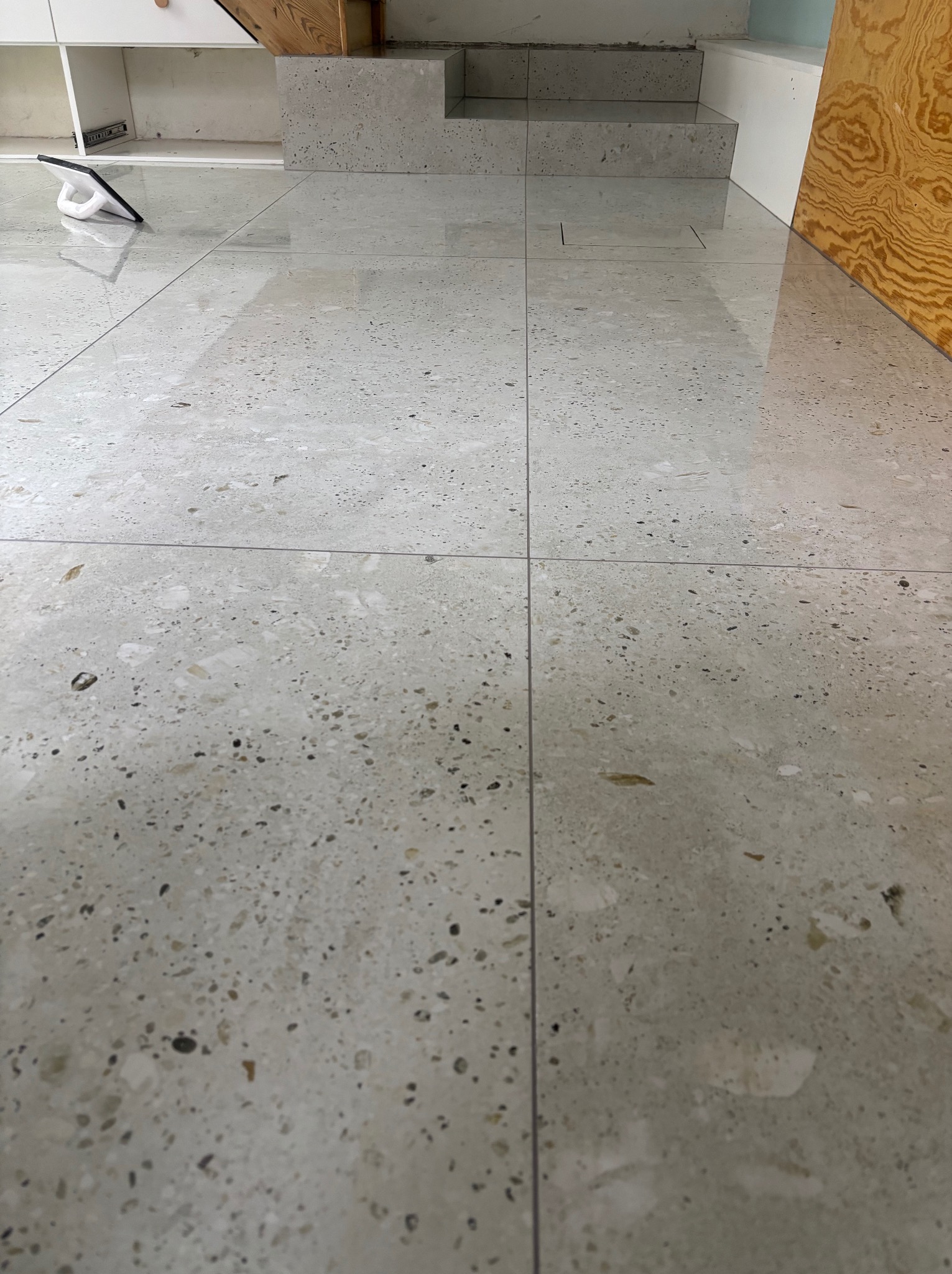 Błyszcząca, jasna posadzka z terazzo, z widocznym fragmentem schodów i narzędziem glazurniczym w tle. Minimalistyczny design, gładka powierzchnia.