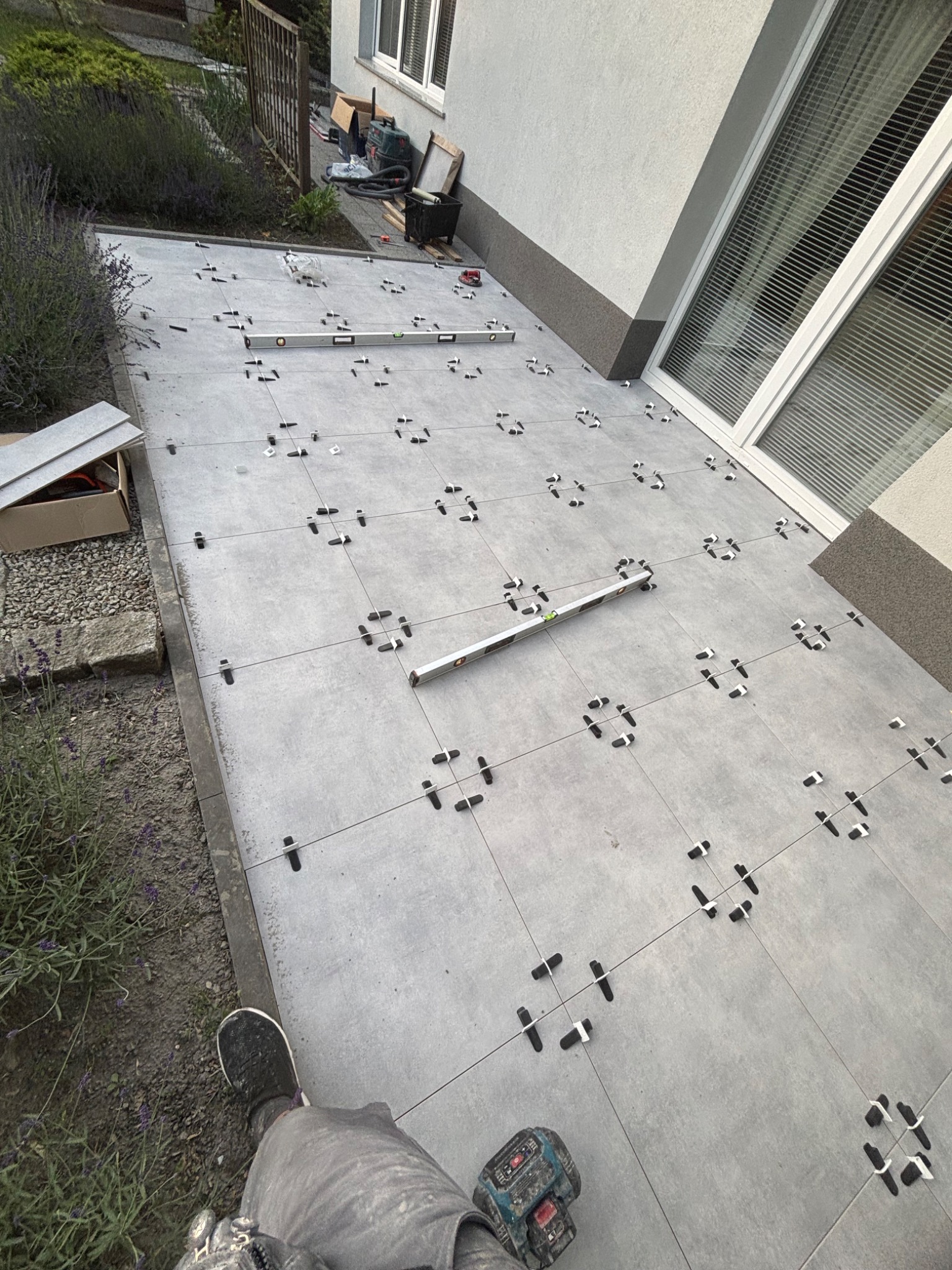 Układanie szarych płytek na zewnątrz budynku z użyciem poziomicy i krzyżyków dystansowych. Widoczne narzędzia i fragment buta glazurnika.