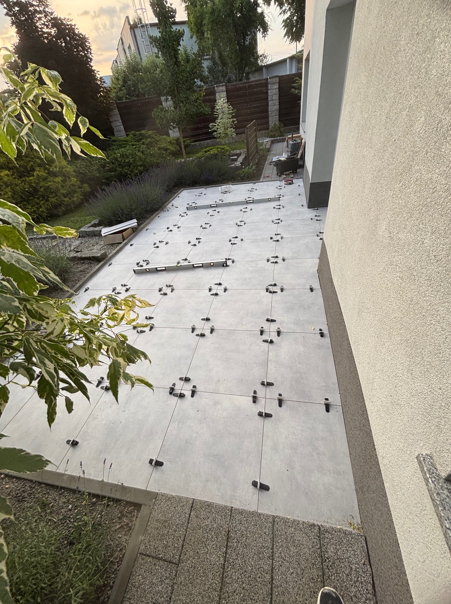 Kładzenie jasnoszarych płytek na tarasie przy domu, z użyciem klipsów poziomujących. Widoczne rośliny, taczka i poziomica. Perspektywa z góry.