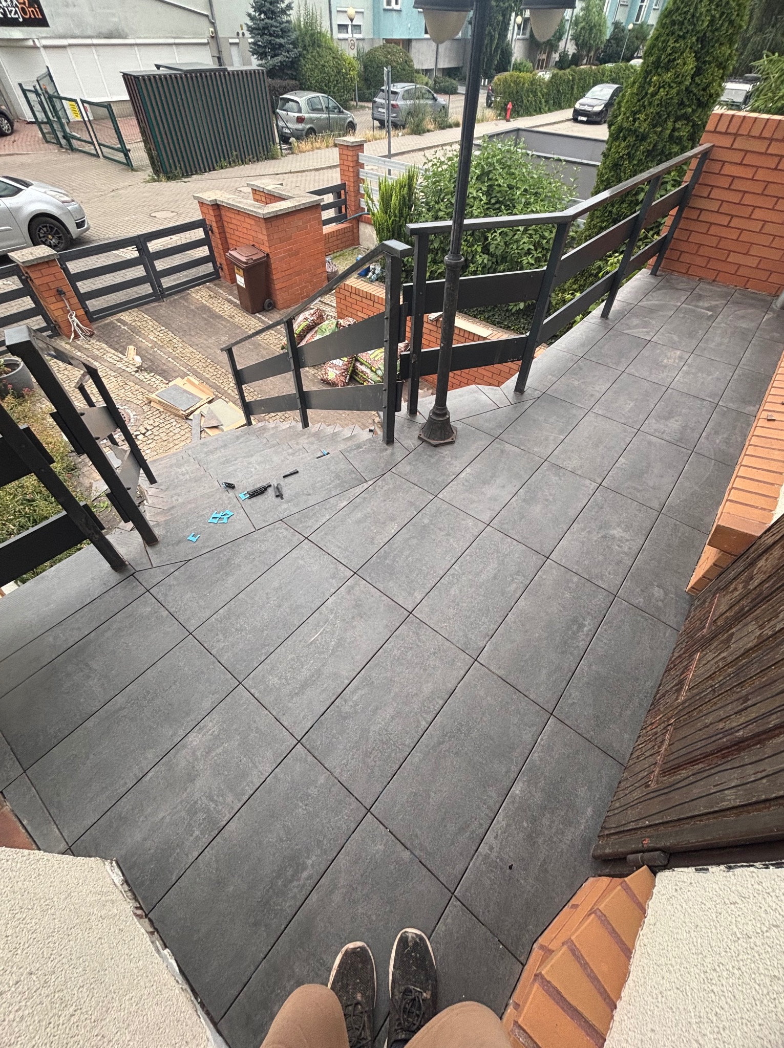 Ułożone grafitowe płytki na schodach zewnętrznych z czarną balustradą. Widoczne narzędzia glazurnicze i fragment butów osoby fotografującej. Tło stanowi zabudowa miejska.