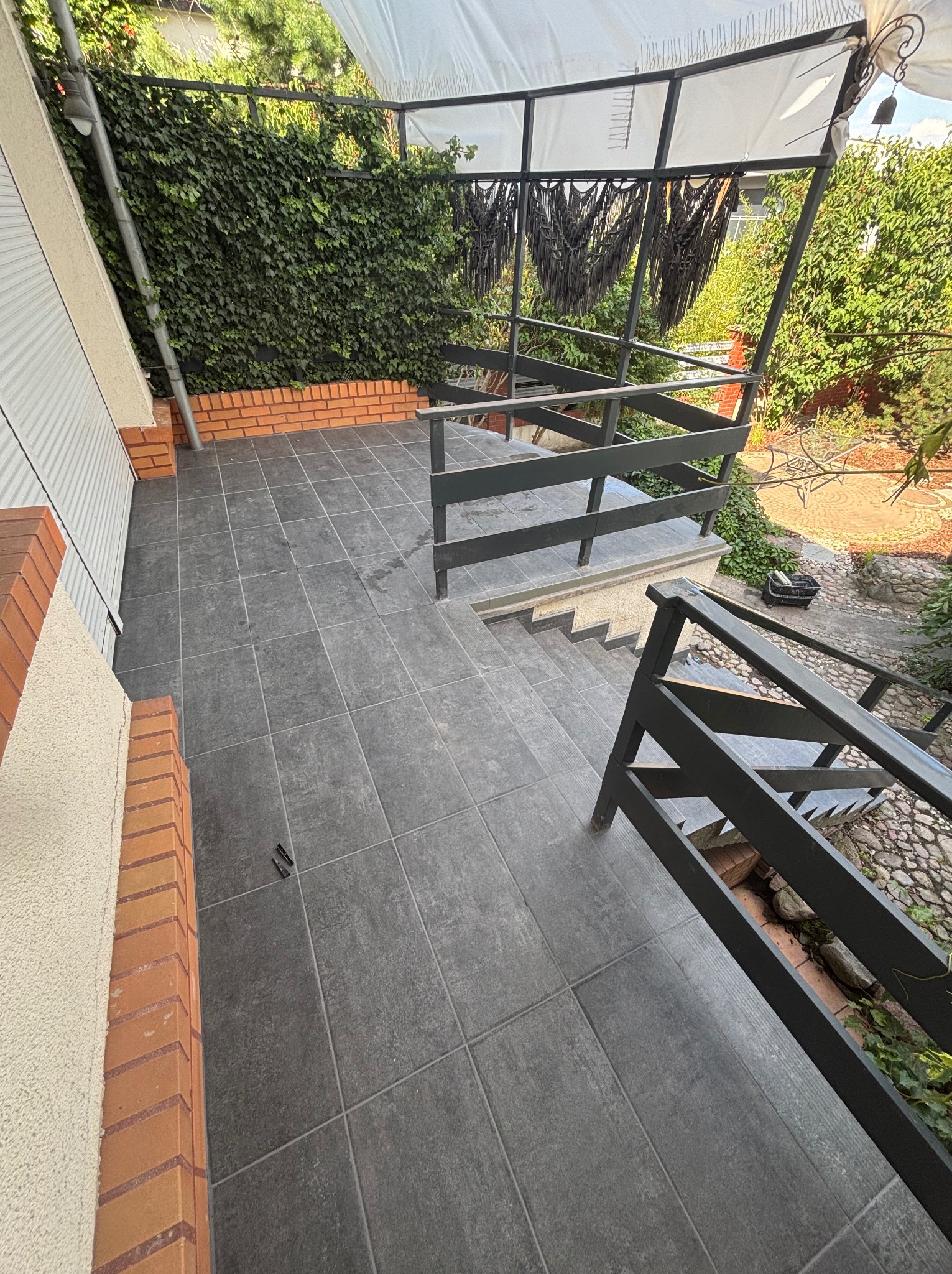 Ułożone szare płytki na tarasie z barierkami i schodami. Ceglana podmurówka, zielona ściana z bluszczu. Widok z góry, ukośne ujęcie.