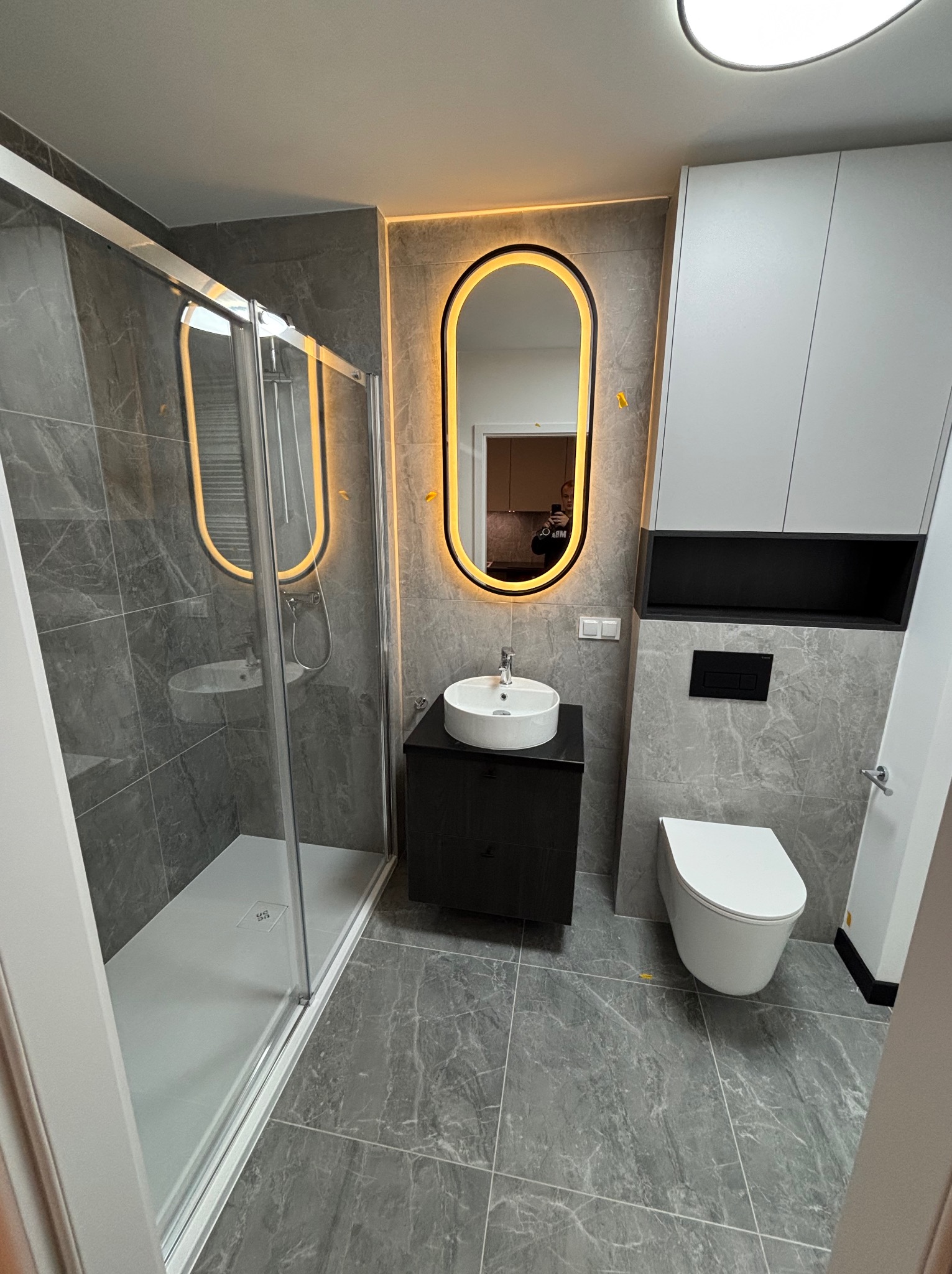 Nowoczesna łazienka z szarymi płytkami imitującymi marmur, podświetlanym lustrem owalnym, kabiną prysznicową i podwieszaną toaletą.