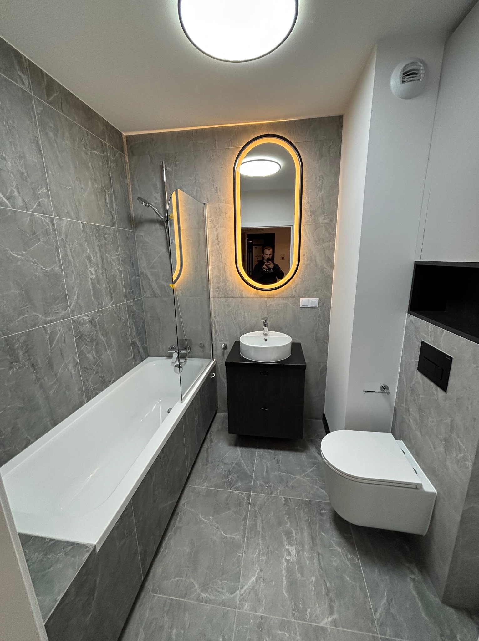 Nowoczesna łazienka w szarościach: wanna z parawanem, podwieszana toaleta i umywalka z podświetlanym lustrem. Marmurowe płytki na ścianach i podłodze.