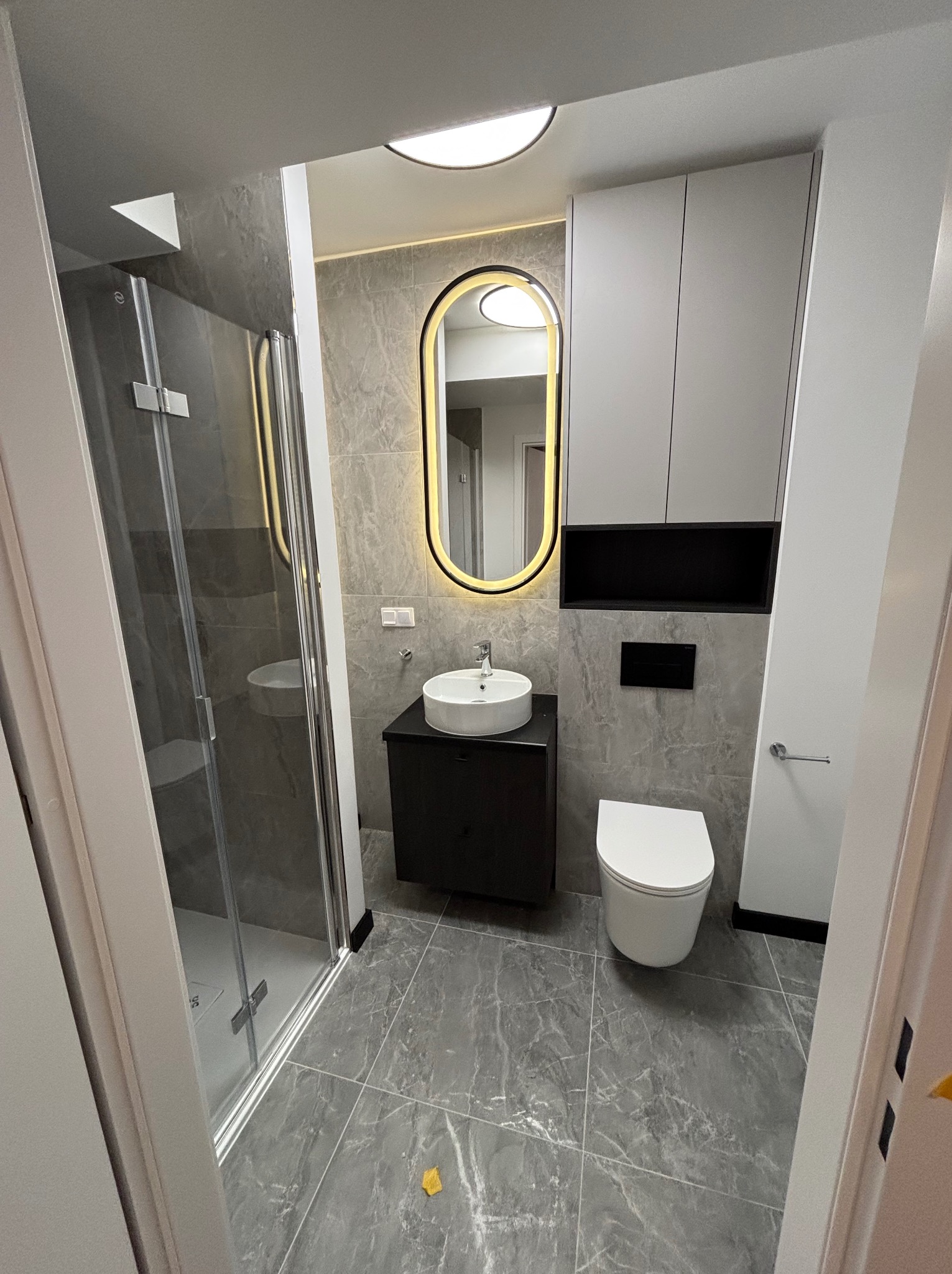 Nowoczesna łazienka z kabiną prysznicową, owalnym lustrem z podświetleniem LED, umywalką nablatową i szarą marmurową płytką. Minimalistyczny design.