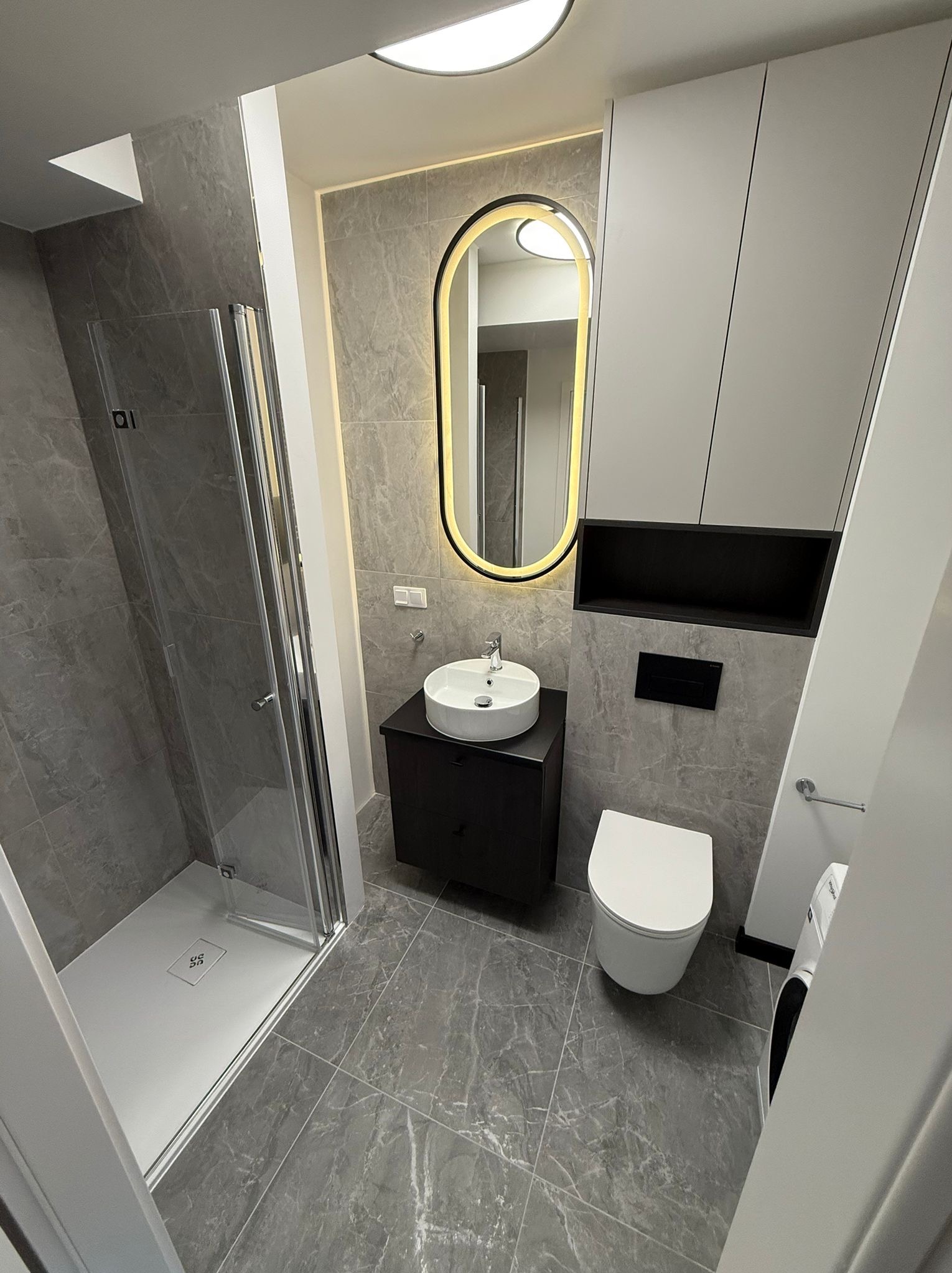 Nowoczesna łazienka w szarościach: kabina prysznicowa, umywalka na szafce, toaleta, podświetlane lustro owalne, płytki imitujące marmur, minimalistyczne szafki.