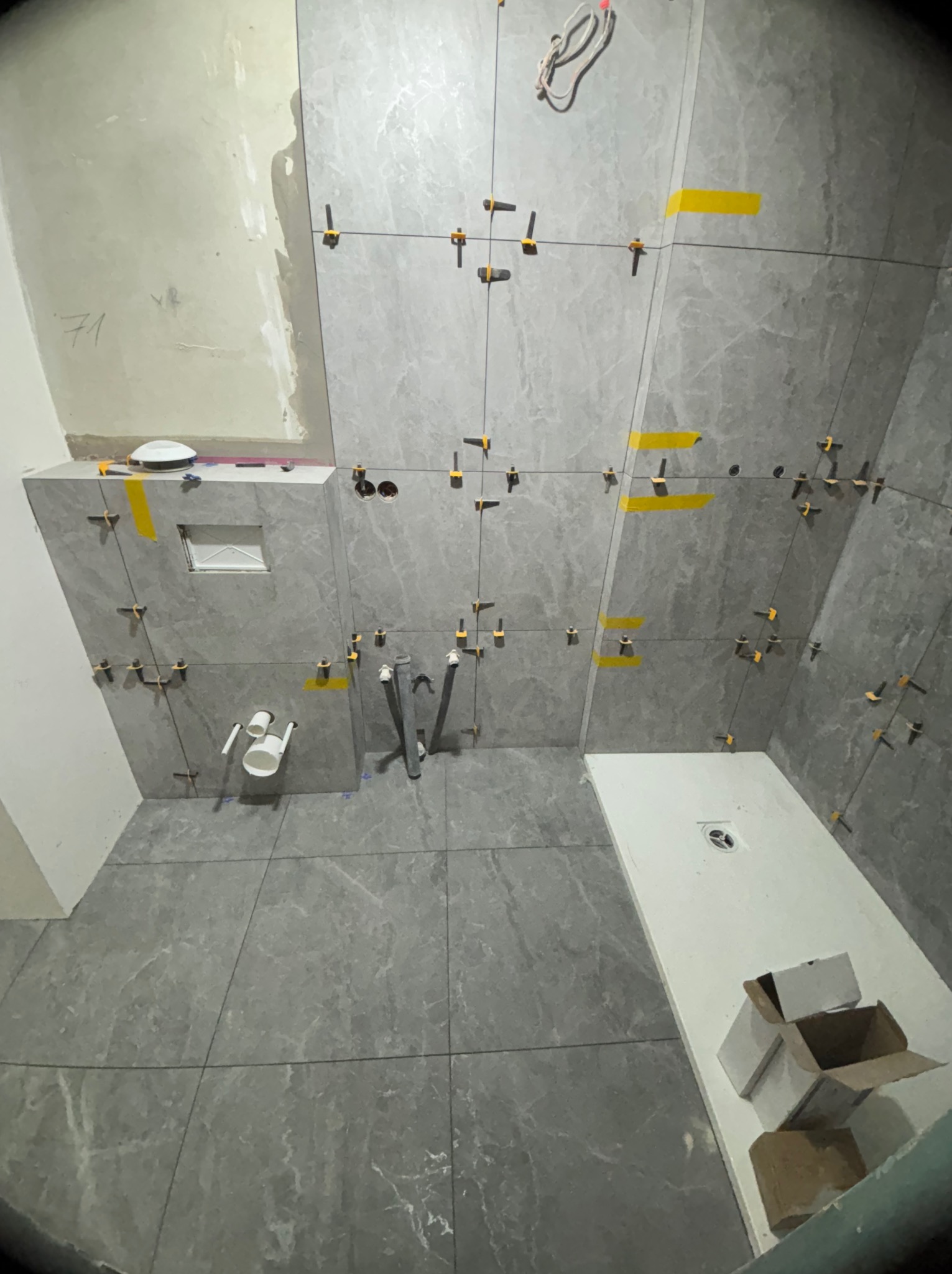 Układanie szarych płytek w łazience, widoczne krzyżyki dystansowe i taśma. Częściowo ułożona podłoga i ściany. W tle elementy instalacji wodnej i odpływ liniowy.