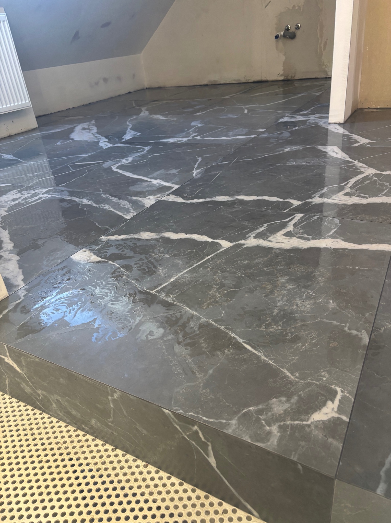 Połyskująca, szara podłoga z marmurowym wzorem w łazience na poddaszu, z widocznymi fragmentami grzejnika i instalacji wodnej. Ukończone płytkowanie.