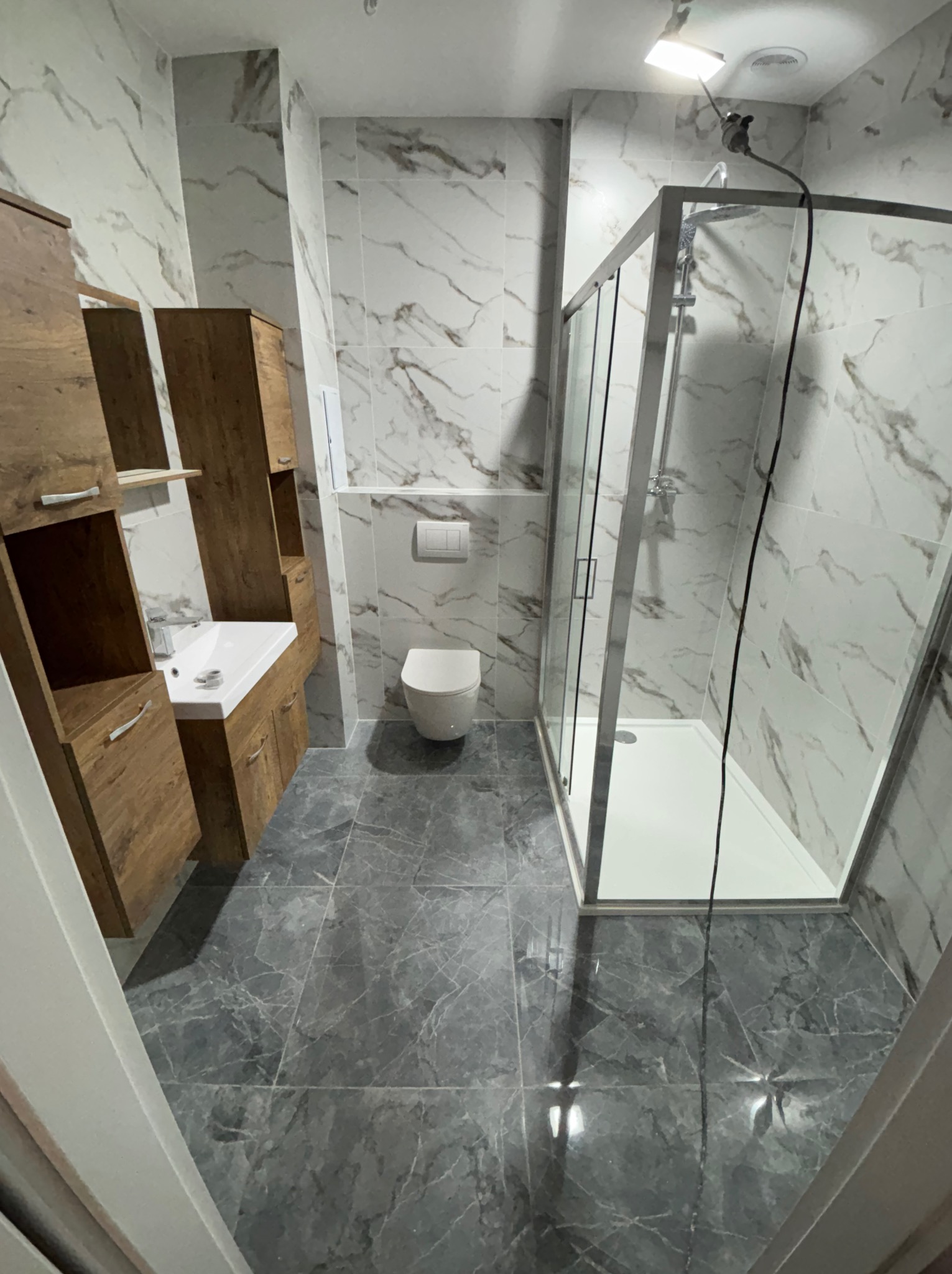 Nowoczesna łazienka z marmurowymi płytkami, drewnianą szafką pod umywalką i kabiną prysznicową. Szare płytki podłogowe z wzorem żyłkowania.