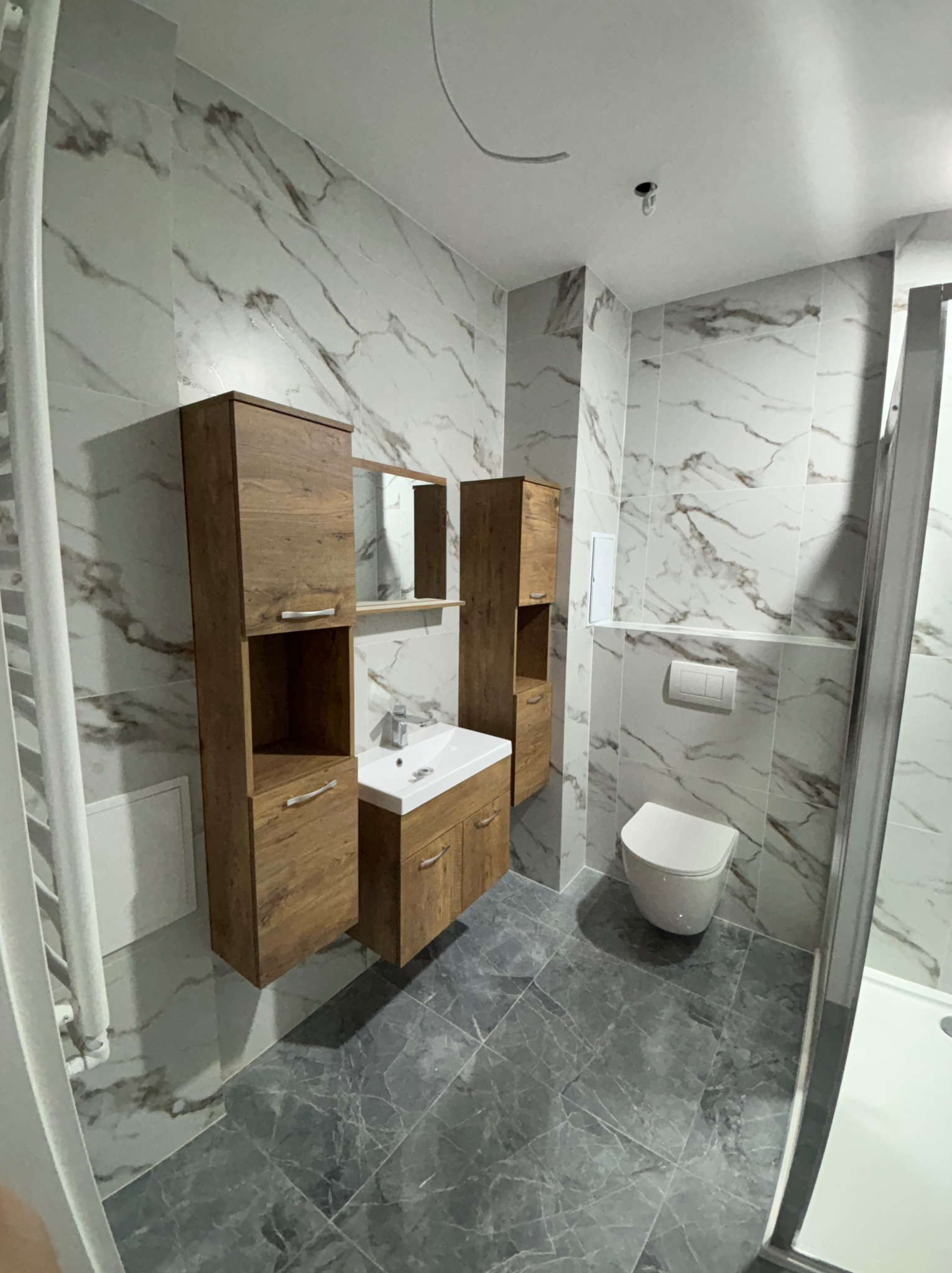 Wykończona łazienka z marmurowymi płytkami, drewnianymi szafkami i szarą podłogą. Widoczna toaleta, umywalka z szafką i kabina prysznicowa.