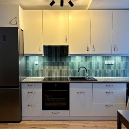 Piotr Kołosowski - Nowoczesna kuchnia z białymi szafkami i srebrną lodówką. Ciemne płytki nad blatem, czarny zlew i płyta indukcyjna. Oświetlenie sufitowe punktowe.