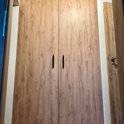 Piotr Kołosowski - Dwudrzwiowa szafa wnękowa z drewnopodobnymi frontami i czarnymi uchwytami, widoczna z perspektywy podłogi, wkomponowana w jasne wnętrze.