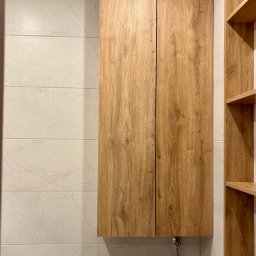 Piotr Kołosowski - Drewniana szafka wisząca nad toaletą w łazience z płytkami imitującymi marmur. Obok półki z drewna w tym samym odcieniu. Ujęcie z bliska.