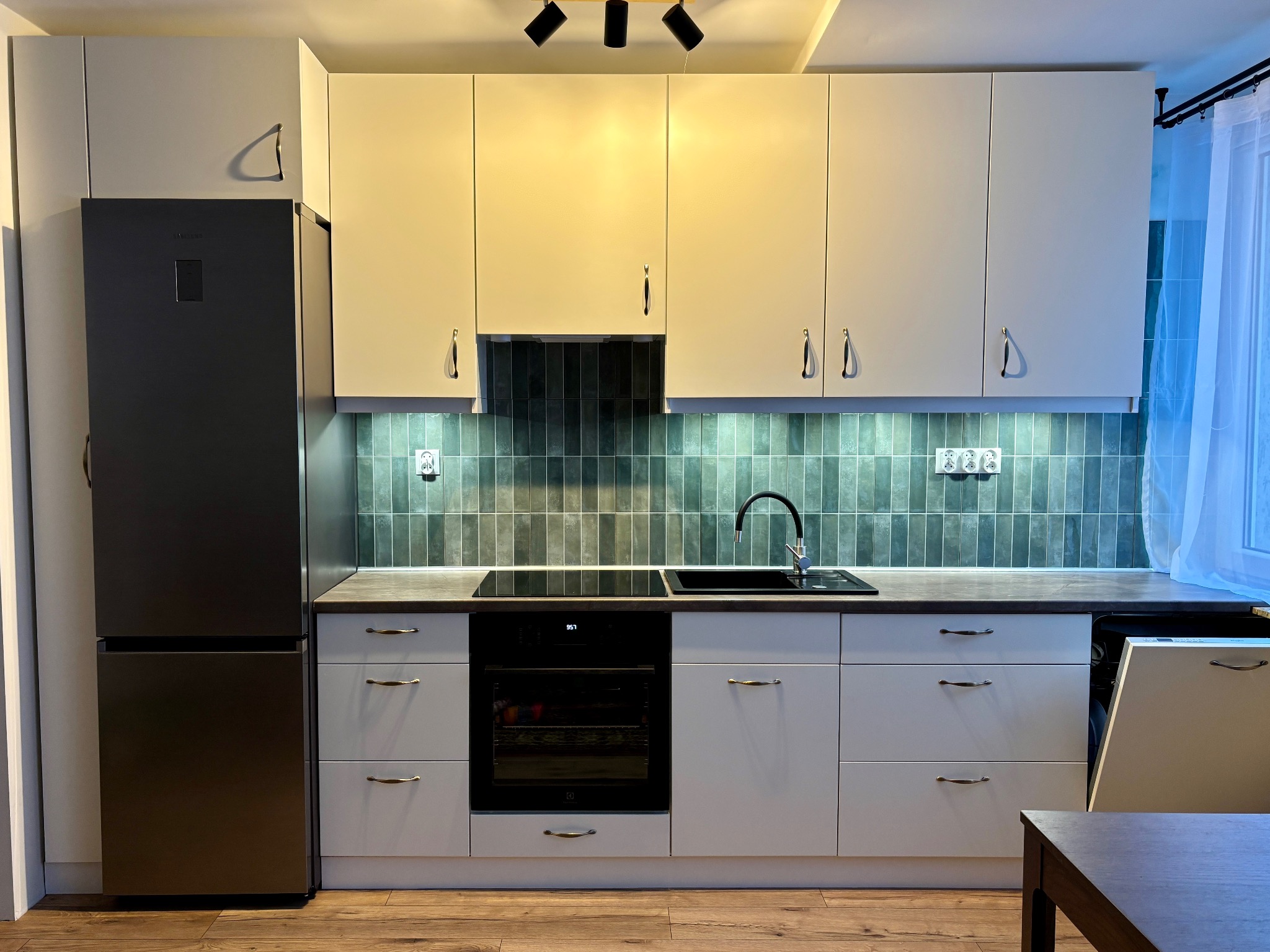 Nowoczesna kuchnia z białymi szafkami i srebrną lodówką. Ciemne płytki nad blatem, czarny zlew i płyta indukcyjna. Oświetlenie sufitowe punktowe.