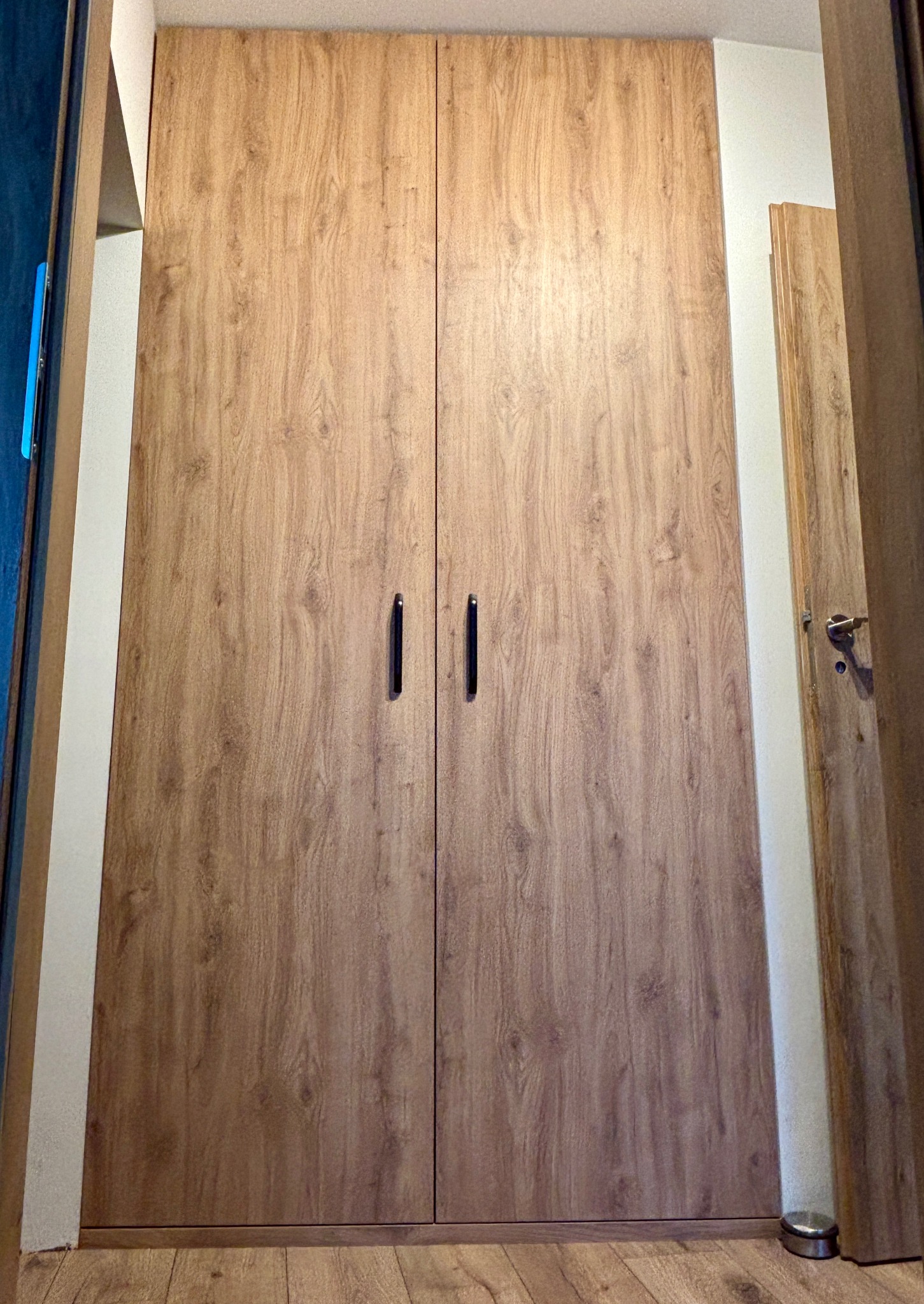 Dwudrzwiowa szafa wnękowa z drewnopodobnymi frontami i czarnymi uchwytami, widoczna z perspektywy podłogi, wkomponowana w jasne wnętrze.