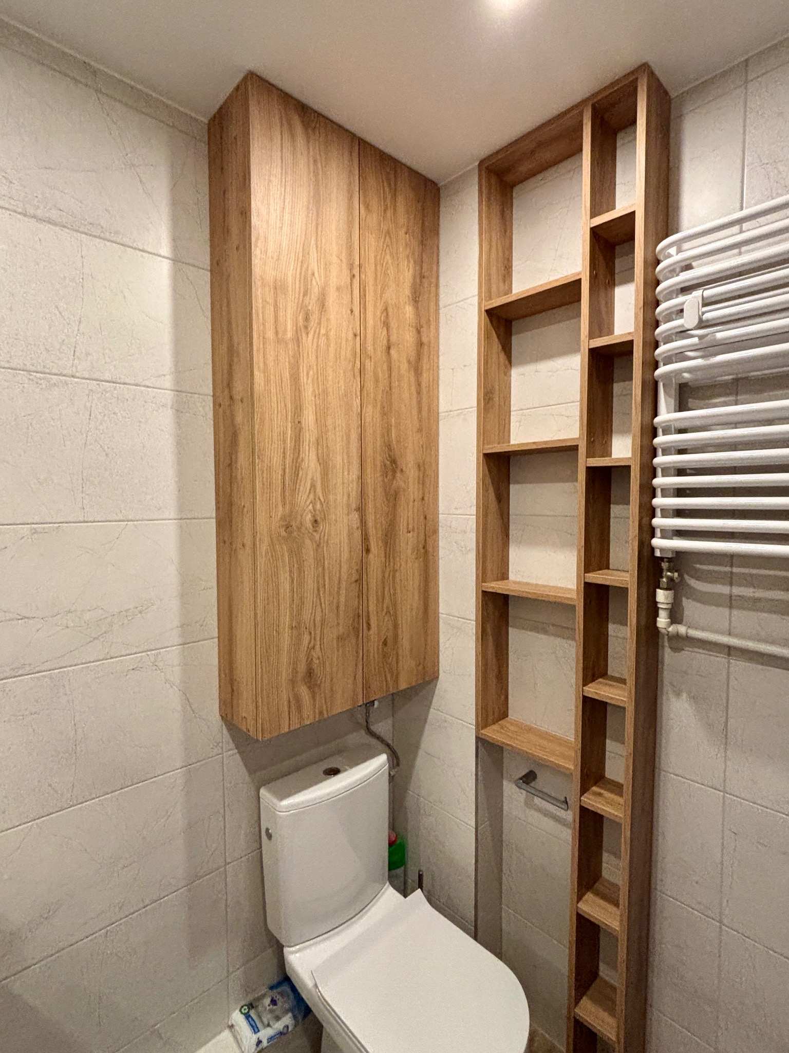 Drewniana szafka wisząca i regał z półkami w łazience nad toaletą, dopasowane kolorystycznie do płytek. Grzejnik drabinkowy po prawej stronie.