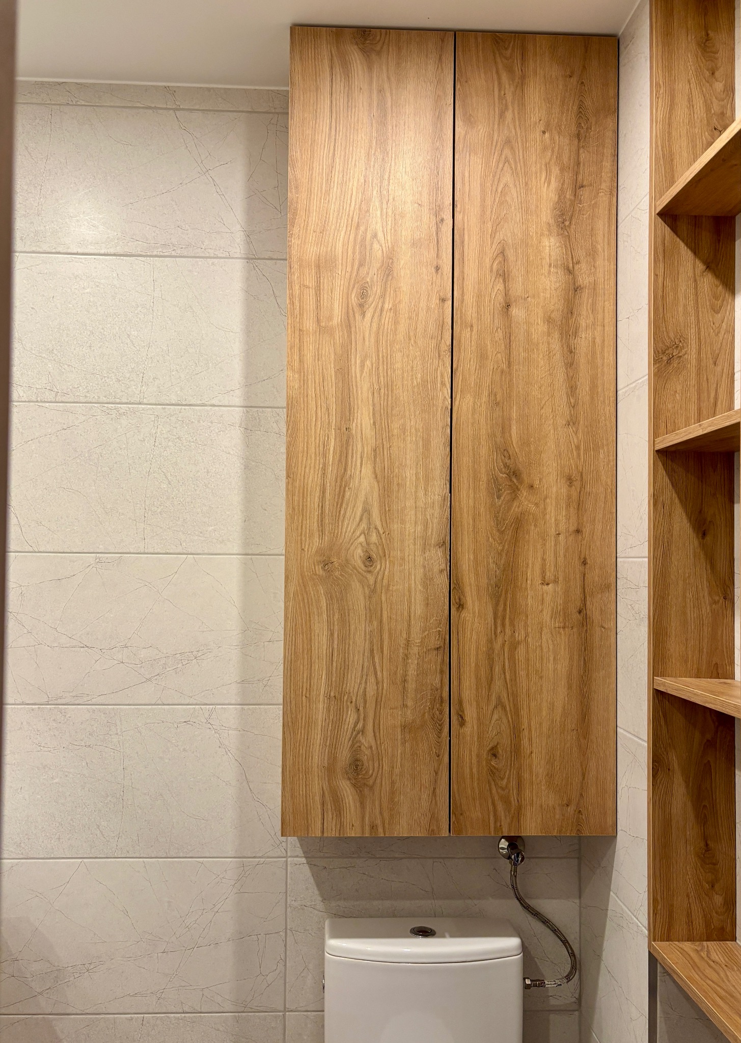 Drewniana szafka wisząca nad toaletą w łazience z płytkami imitującymi marmur. Obok półki z drewna w tym samym odcieniu. Ujęcie z bliska.