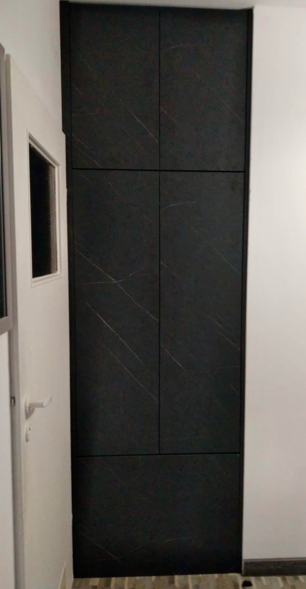 Czarna szafa wnękowa z subtelnym wzorem marmuru, wkomponowana w wąskie pomieszczenie, widoczna obok białych drzwi.