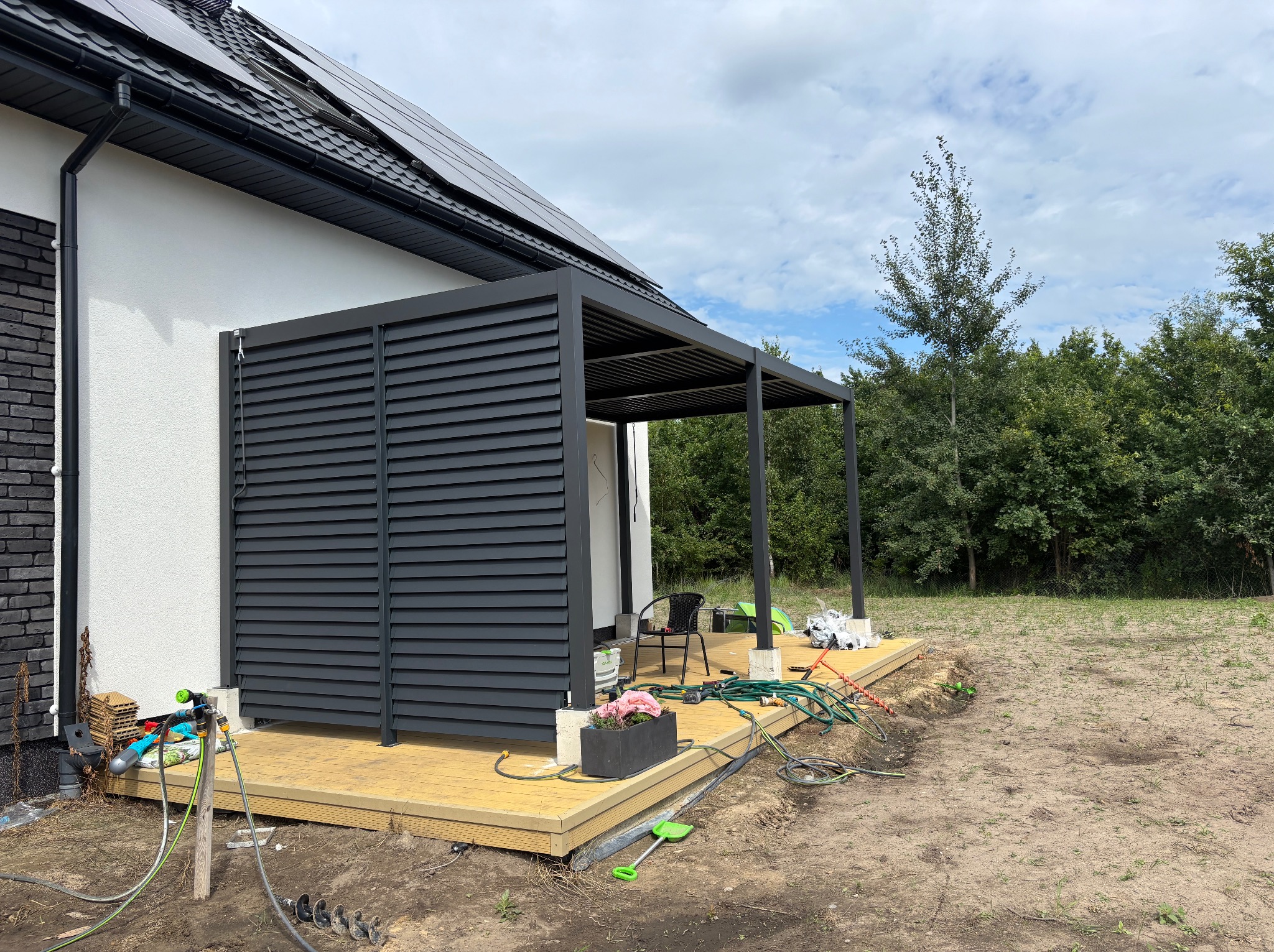 Nowoczesna pergola metalowa z żaluzjową ścianą boczną, połączona z drewnianym tarasem przy domu z panelami solarnymi na dachu. Widoczne akcesoria ogrodowe.