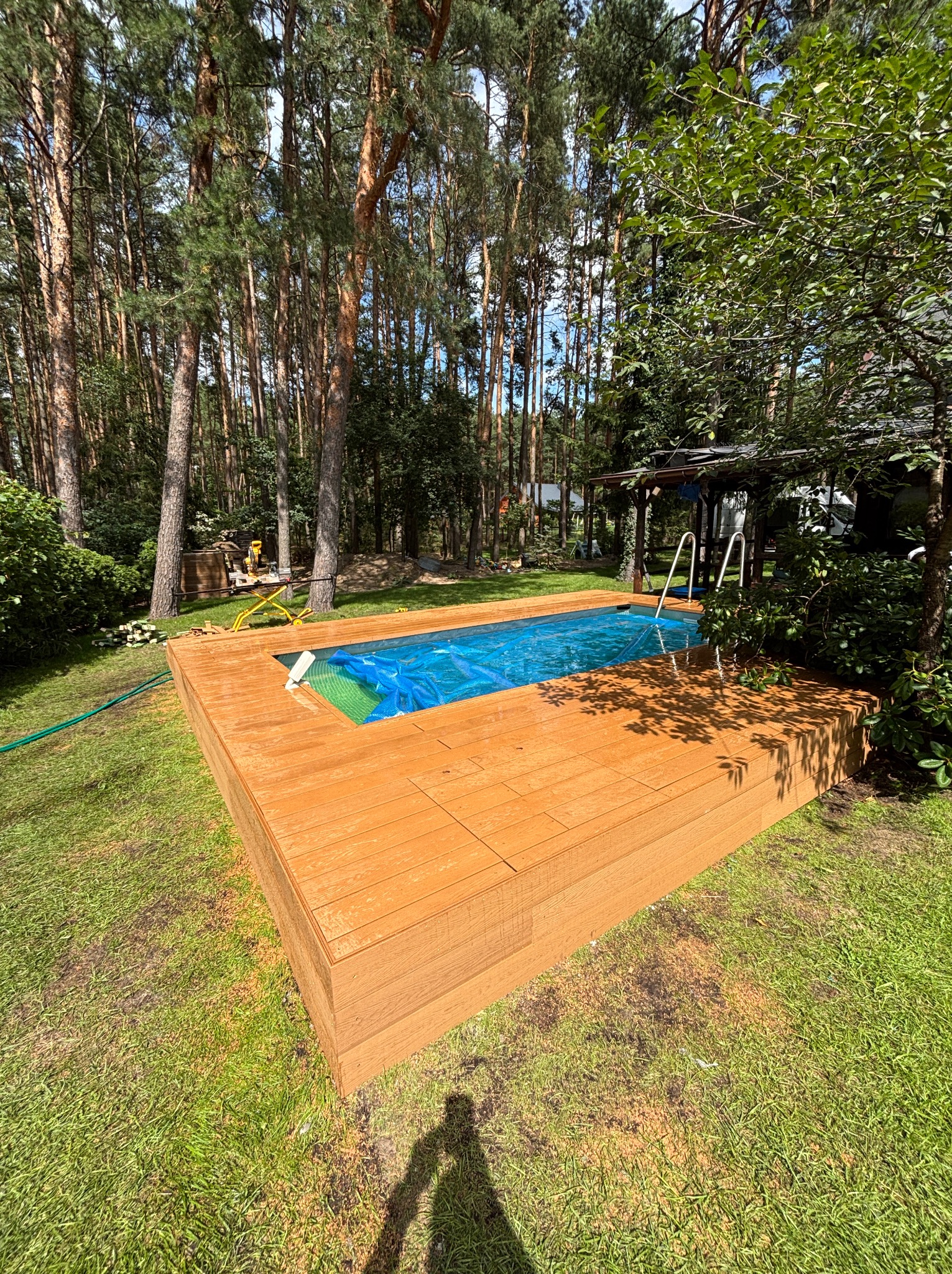 Drewniany taras okalający basen w ogrodzie leśnym, z widoczną drabinką i niebieską plandeką na wodzie. Świeżo położona deska tarasowa w ciepłym odcieniu brązu.