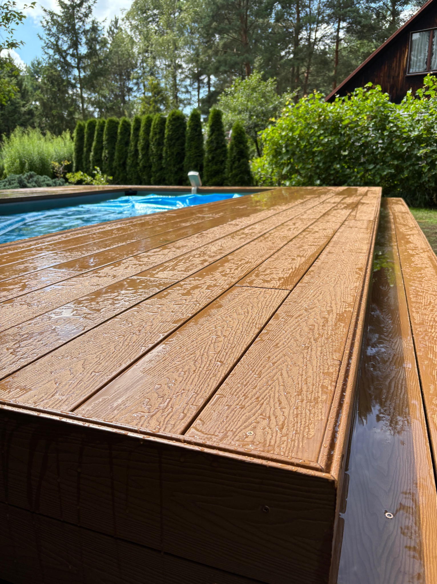 Wodoodporny taras kompozytowy przy basenie, deski w odcieniach brązu, widoczne krople wody. Tło: zielone tuje i fragment domu z ciemnego drewna.