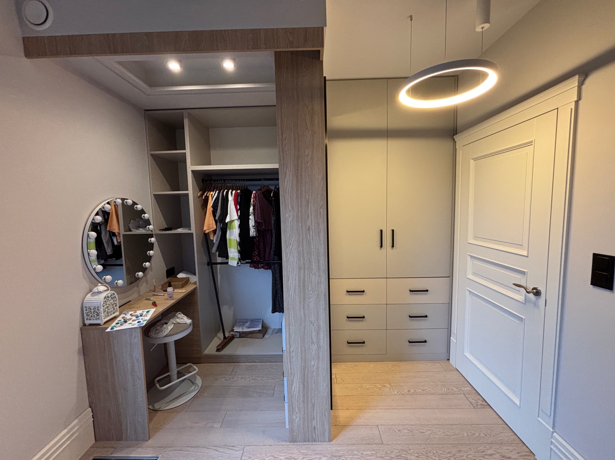 Nowoczesna garderoba z szafą wnękową, toaletką z lustrem i krzesłem. Jasne drewno i szarość tworzą spójną całość, oświetloną designerską lampą.