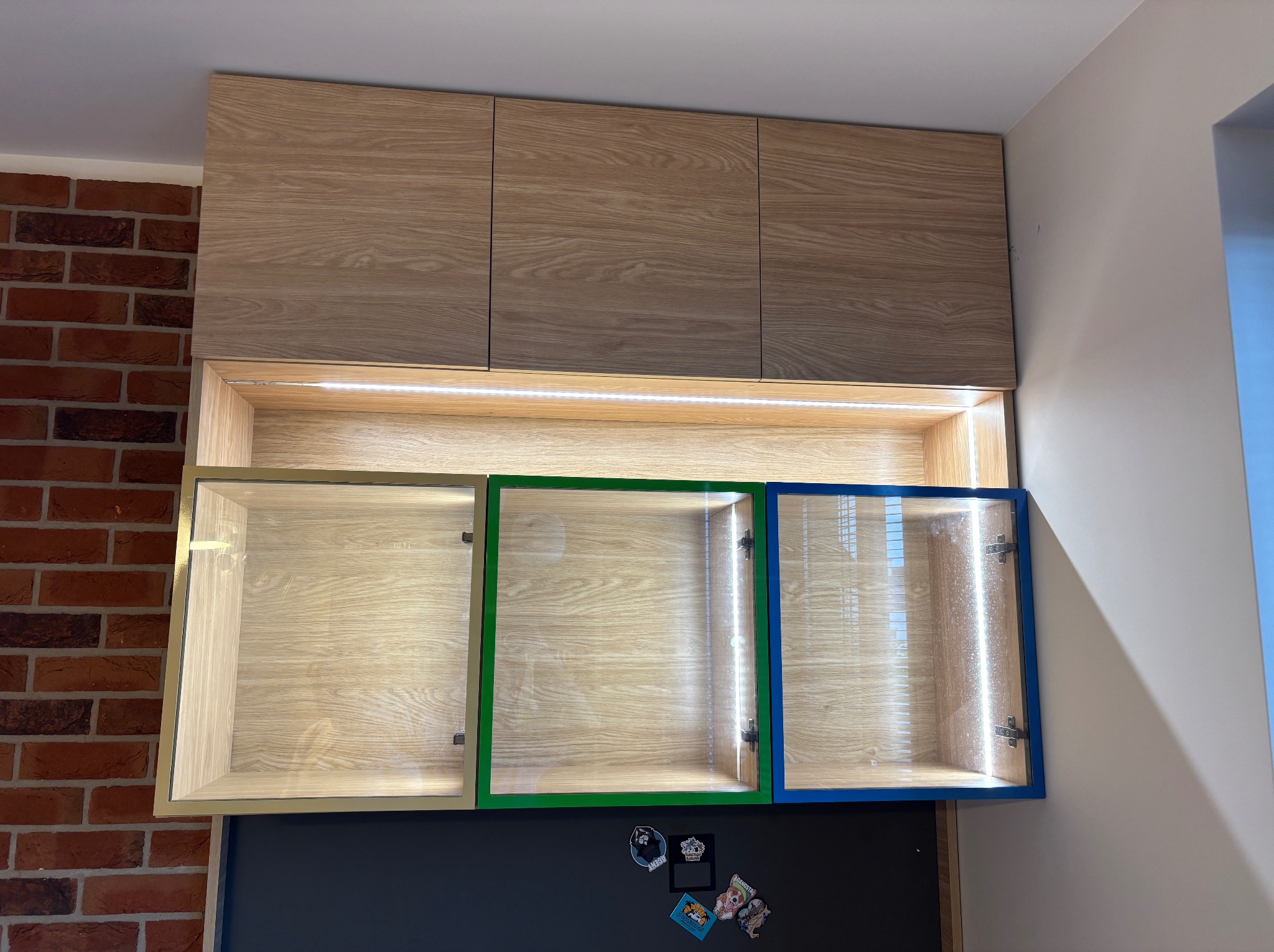 Nowoczesne szafki wiszące z oświetleniem LED, fronty z kolorowymi ramkami (złota, zielona, niebieska). Na dole czarna tablica z magnesami. Ceglana ściana po lewej.