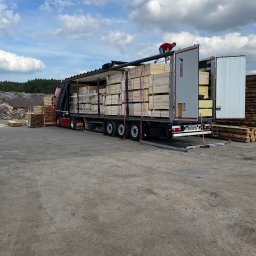 Zakład Stolarski M.L.Malinowscy s.j. - Ciężarówka z otwartą naczepą wypełniona pakietami tarcicy budowlanej. Na naczepie stoi mężczyzna, nadzorując rozładunek. W tle składowane drewno.