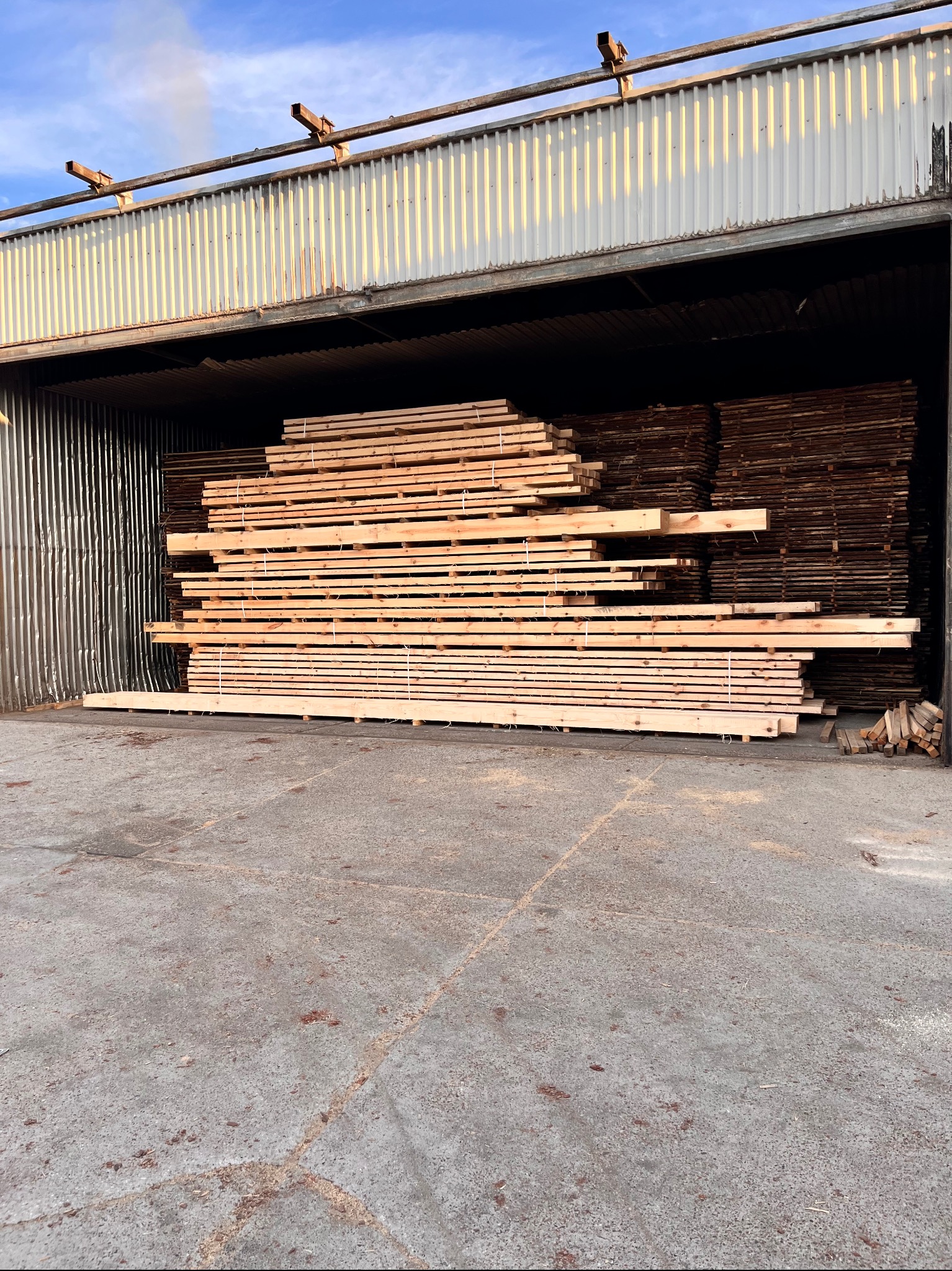 Stos tarcicy budowlanej w magazynie z metalową konstrukcją. Jasne, świeżo przetarte deski ułożone w równe warstwy, zabezpieczone sznurkiem. W tle ciemniejsze drewno.