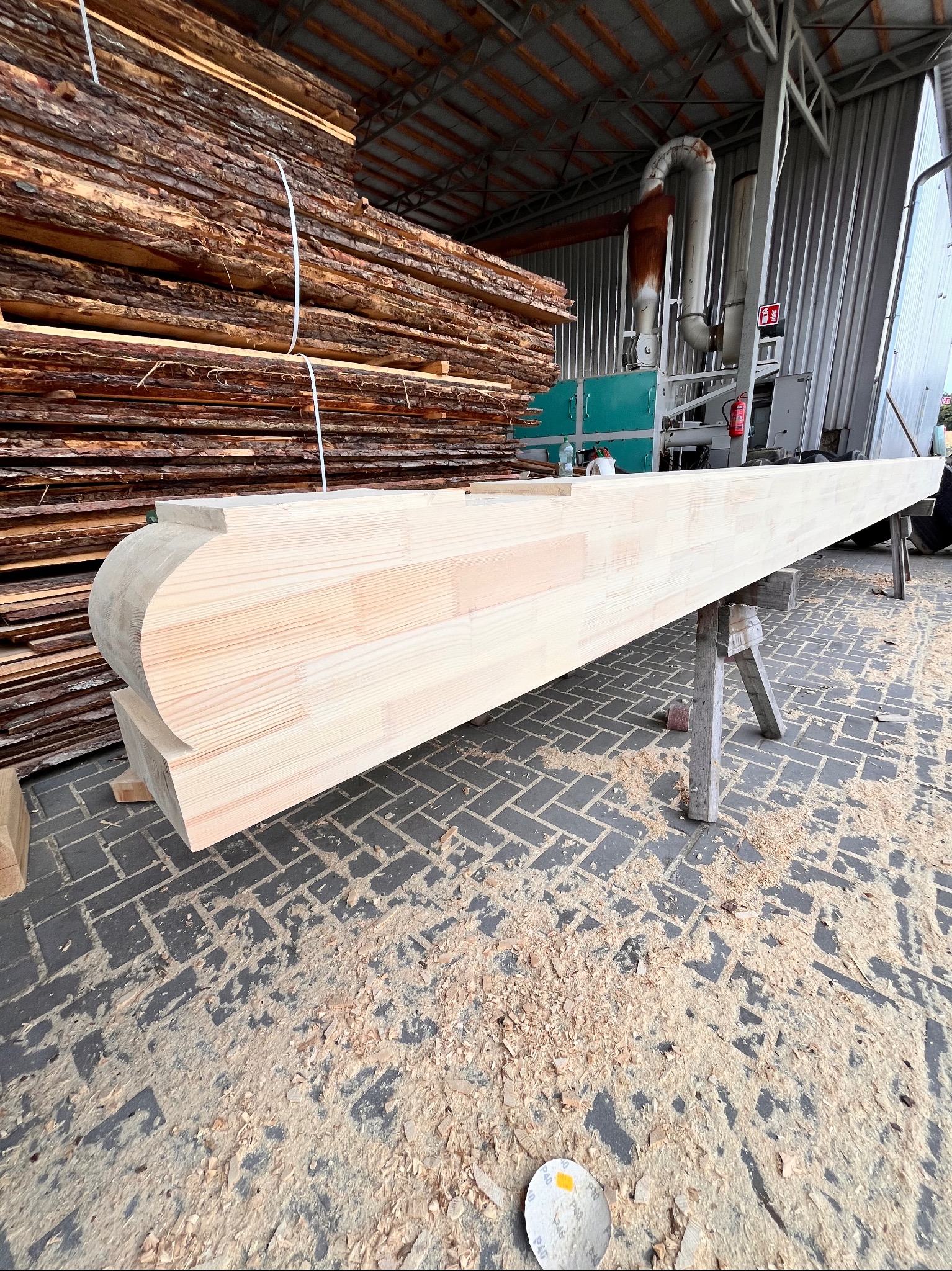 Krawędziak z zaokrąglonym zakończeniem, ułożony na kozłach w tartaku. W tle stosy tarcicy i maszyny do obróbki drewna. Wióry na posadzce.