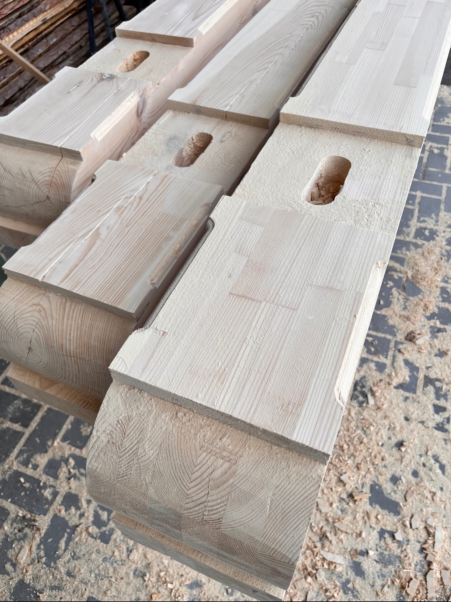 Obróbka drewna: Jasne, heblowane belki z wyfrezowanymi otworami, ułożone równolegle. Widoczne ślady obróbki i wióry, ukazujące etap przygotowania konstrukcji.