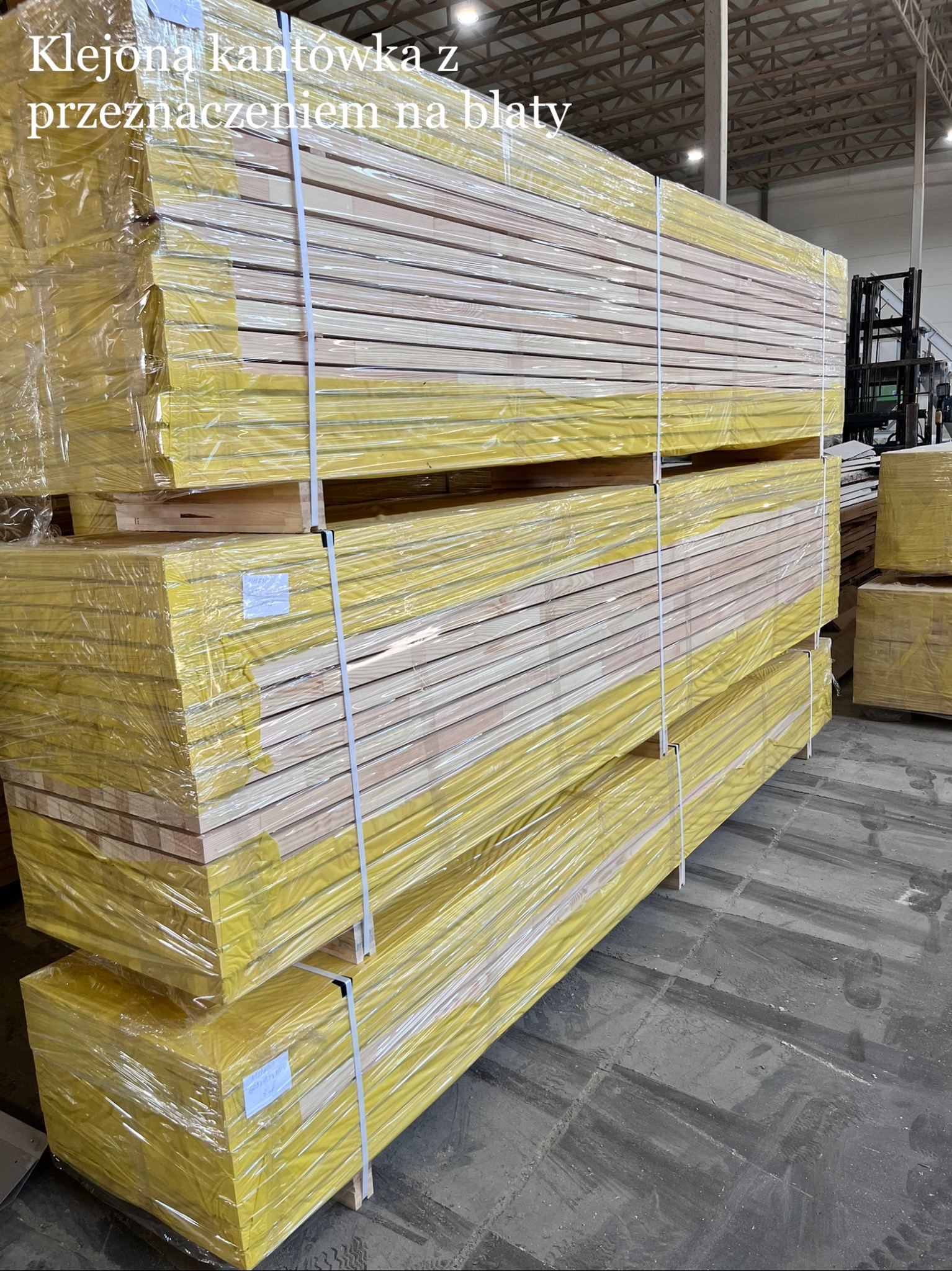 Stos kantówki klejonej, owinięty żółtą folią, przeznaczonej na blaty, w magazynie z wózkiem widłowym w tle. Widoczne warstwy drewna i zabezpieczenia.