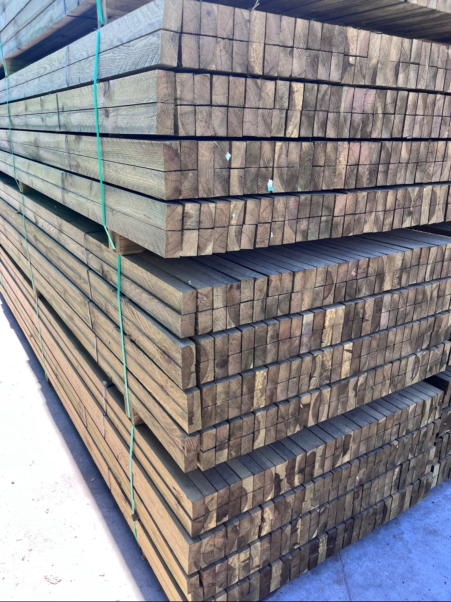 Stos impregnowanej tarcicy budowlanej o przekroju kwadratowym, spiętej zielonymi taśmami, ułożonej na betonowym podłożu.