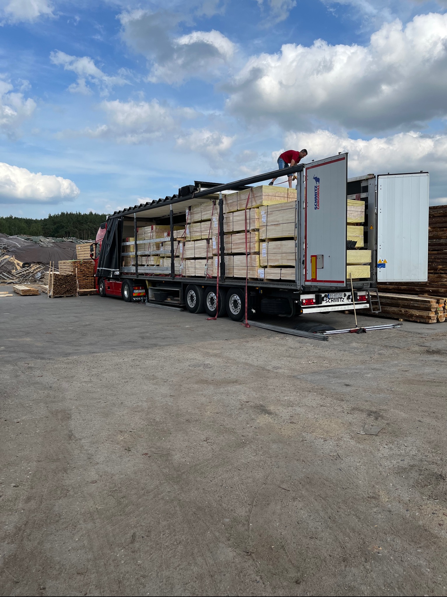 Ciężarówka z otwartą naczepą wypełniona pakietami tarcicy budowlanej. Na naczepie stoi mężczyzna, nadzorując rozładunek. W tle składowane drewno.