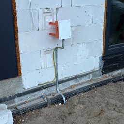 SŁAWOMIR RUDNIK BSM-SERWIS - Pomarańczowo-biała skrzynka elektryczna na ścianie z białych bloczków, z widocznym uziemieniem i rurą ochronną, na tle fragmentu okna i drzwi.