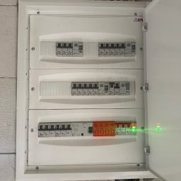 SŁAWOMIR RUDNIK BSM-SERWIS - Rozdzielnica elektryczna ETI z bezpiecznikami i włącznikami, widoczne przewody i fragment ściany. Otwarta obudowa, świecące diody sygnalizacyjne.
