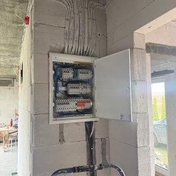 SŁAWOMIR RUDNIK BSM-SERWIS - Otwarta skrzynka elektryczna z licznymi przewodami na ścianie z betonu komórkowego w surowym wnętrzu budynku w trakcie budowy. Widoczne zabezpieczenia i instalacje.