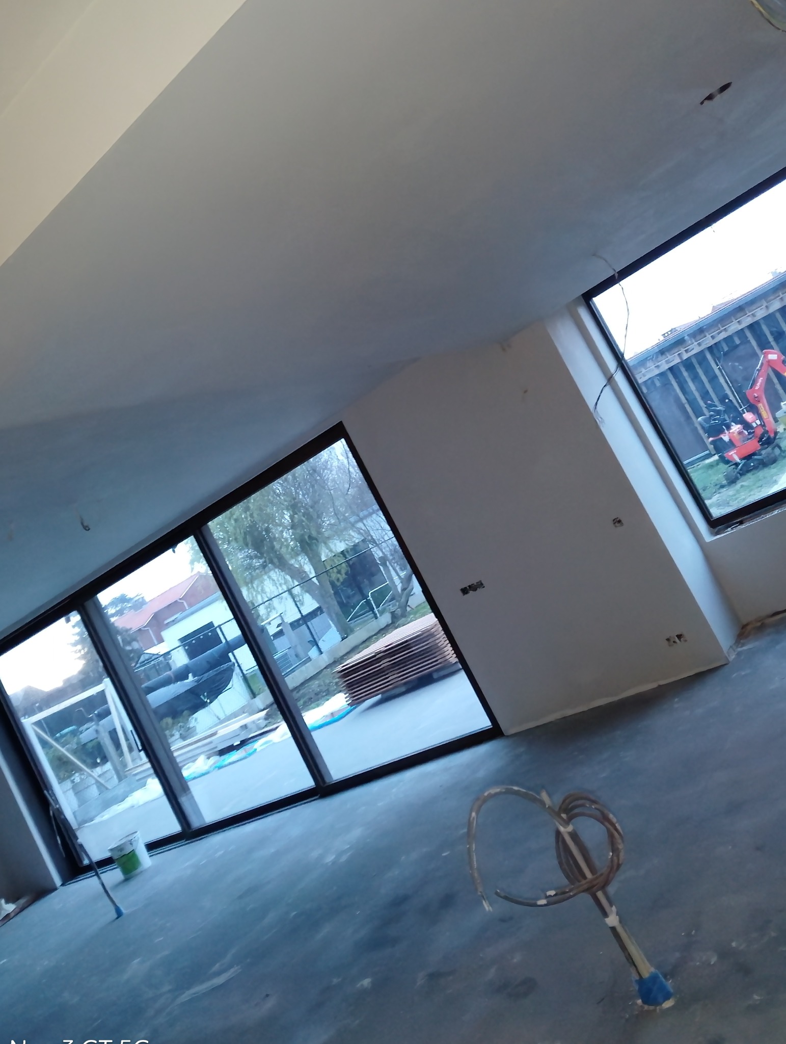 Wnętrze po szpachlowaniu ścian i sufitu, widoczne duże okna z widokiem na zewnątrz, na podłodze leży mieszadło do zaprawy. Ściany przygotowane do dalszych prac.