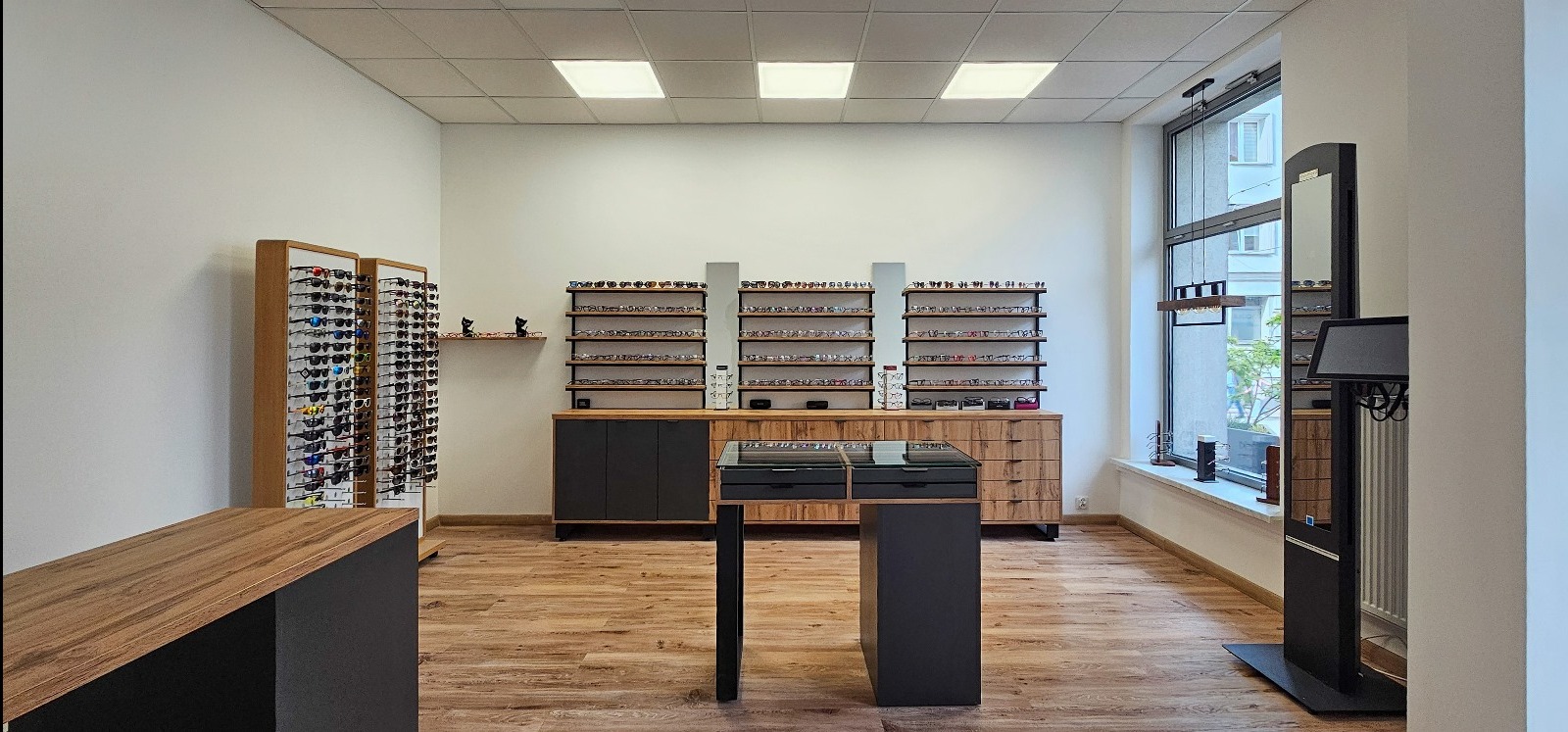 Jasne wnętrze salonu optycznego z ekspozycją okularów na półkach i w gablotach. Widoczne drewniane podłogi i nowoczesne oświetlenie sufitowe. Duże okno z widokiem na zewnątrz.