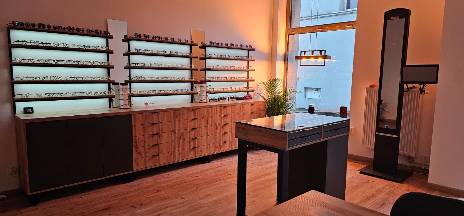 Salon optyczny z ekspozycją okularów na podświetlanych półkach, drewniane meble i stanowisko obsługi klienta w nowoczesnym wnętrzu.