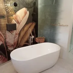 Nowoczesna, wolnostojąca wanna w łazience z motywem roślinnym na ścianie i dekoracyjnym oświetleniem. Widoczna bateria wannowa i koszyk na akcesoria.