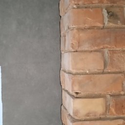 AnDarBud Dariusz Zieliński - Fragment ściany z surowym tynkiem sąsiadującym z dekoracyjną ścianą z czerwonej cegły, ukazujący kontrast faktur i materiałów wykończeniowych.