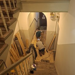 AnDarBud Dariusz Zieliński - Dwóch mężczyzn montuje drewnianą podłogę na klatce schodowej w kamienicy. Widok z góry na etap prac. Widoczne narzędzia i materiały budowlane.
