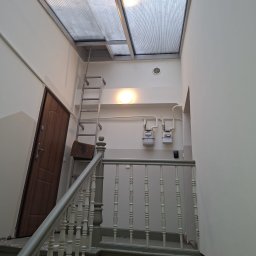 AnDarBud Dariusz Zieliński - Klatka schodowa w trakcie prac wykończeniowych: drewniana balustrada, drabina oparta o ścianę, widoczne liczniki gazu i przeszklony sufit.