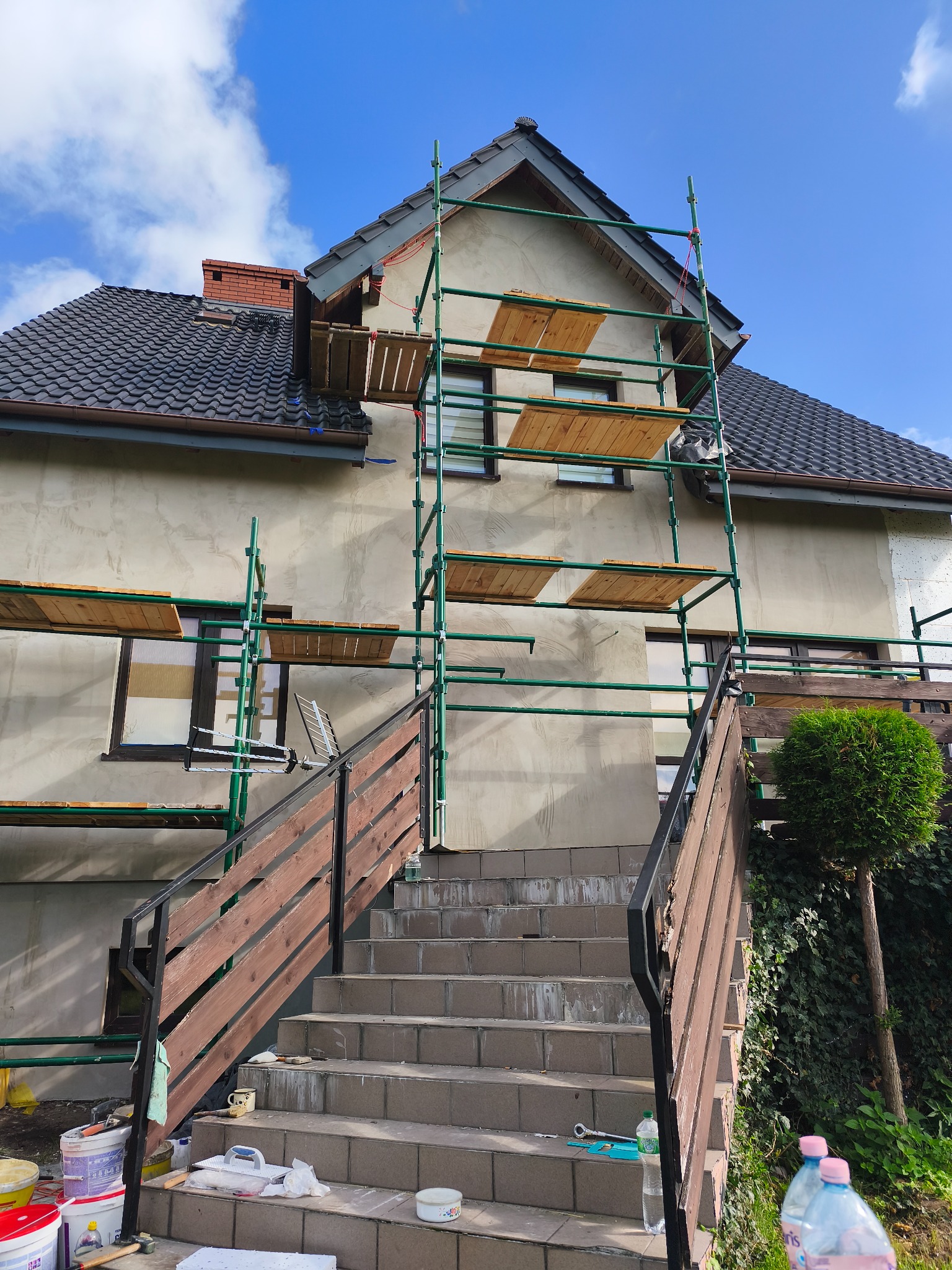 Dom w trakcie ocieplania, z widocznymi rusztowaniami i fragmentem ocieplonej ściany. Na schodach widoczne narzędzia i materiały budowlane.