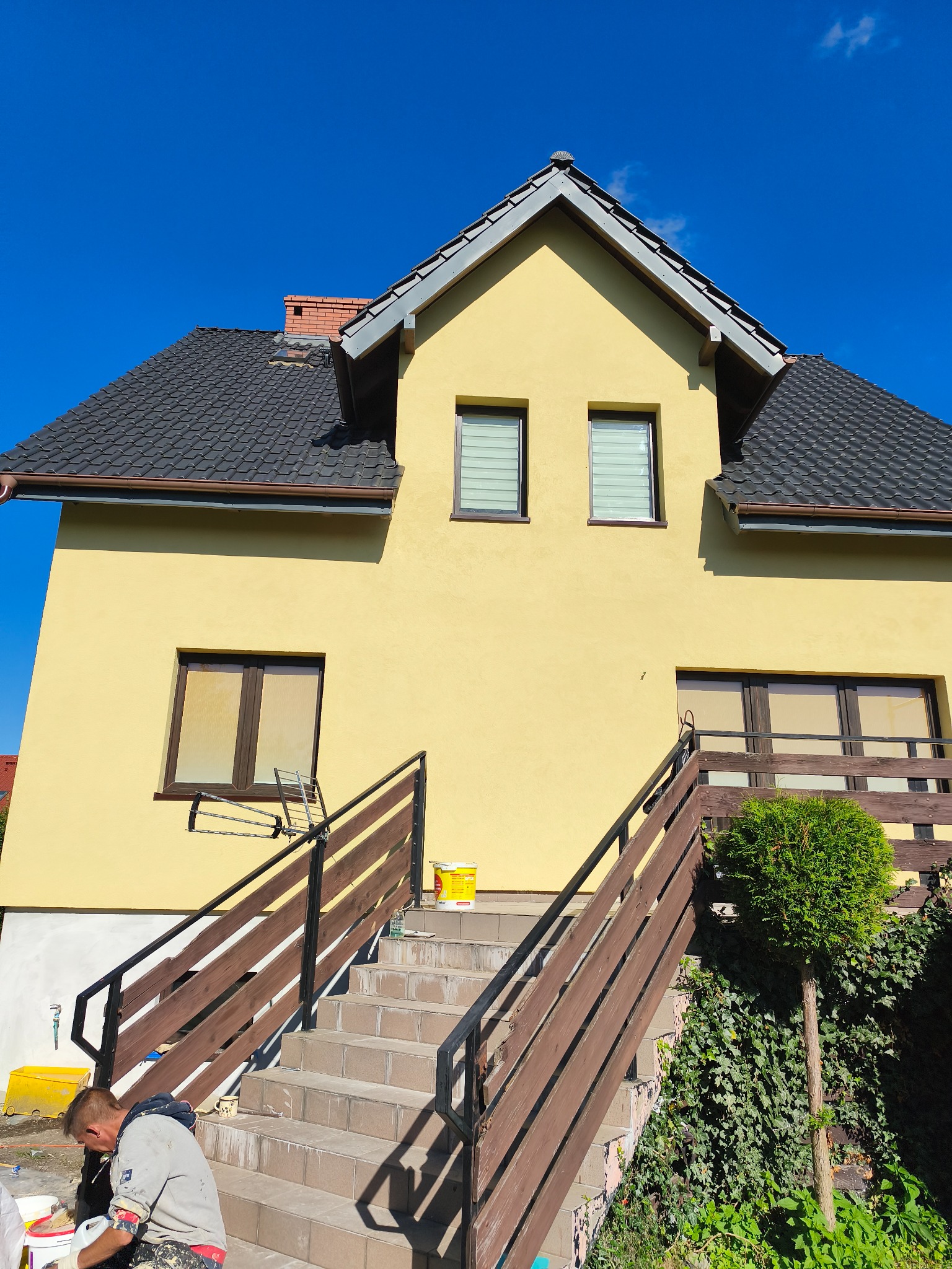 Żółta elewacja domu jednorodzinnego po malowaniu w Gdańsku, widoczne okna z roletami i schody wejściowe. Pracownik w trakcie prac wykończeniowych.