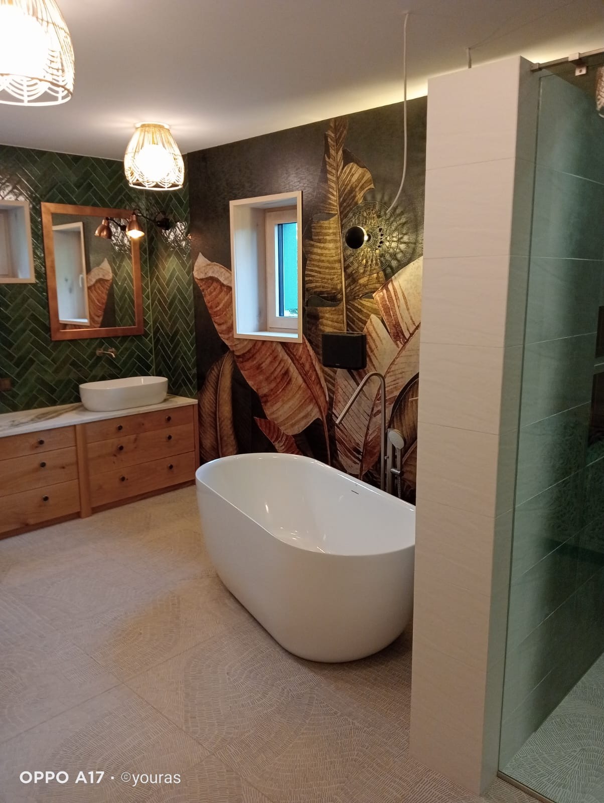 Nowoczesna łazienka z zielonymi płytkami ułożonymi w jodełkę, wolnostojącą wanną i szafką z umywalką. Na ścianie dekoracja z motywem liści.