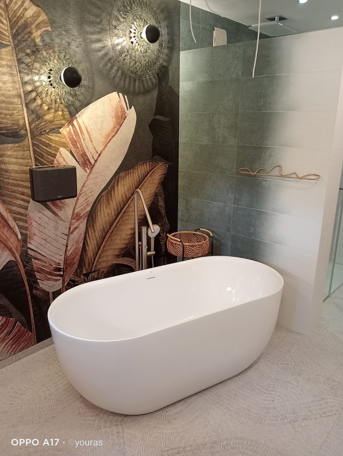 Nowoczesna, wolnostojąca wanna w łazience z motywem roślinnym na ścianie i dekoracyjnym oświetleniem. Widoczna bateria wannowa i koszyk na akcesoria.